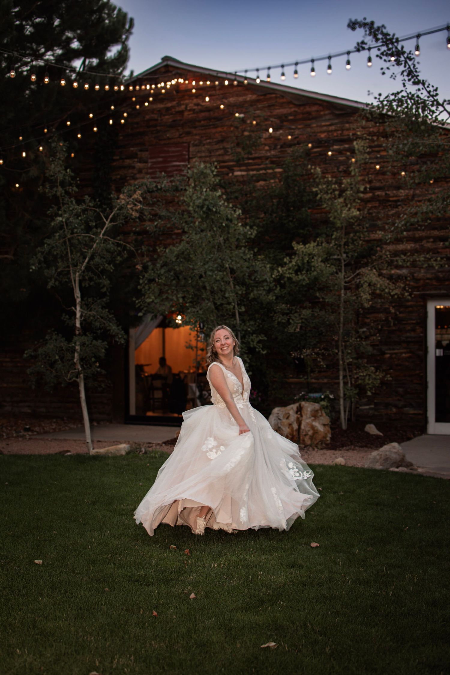 Bride twirling in a flowing white wedding dress on green lawn at dusk with string lights and rustic brick venue.