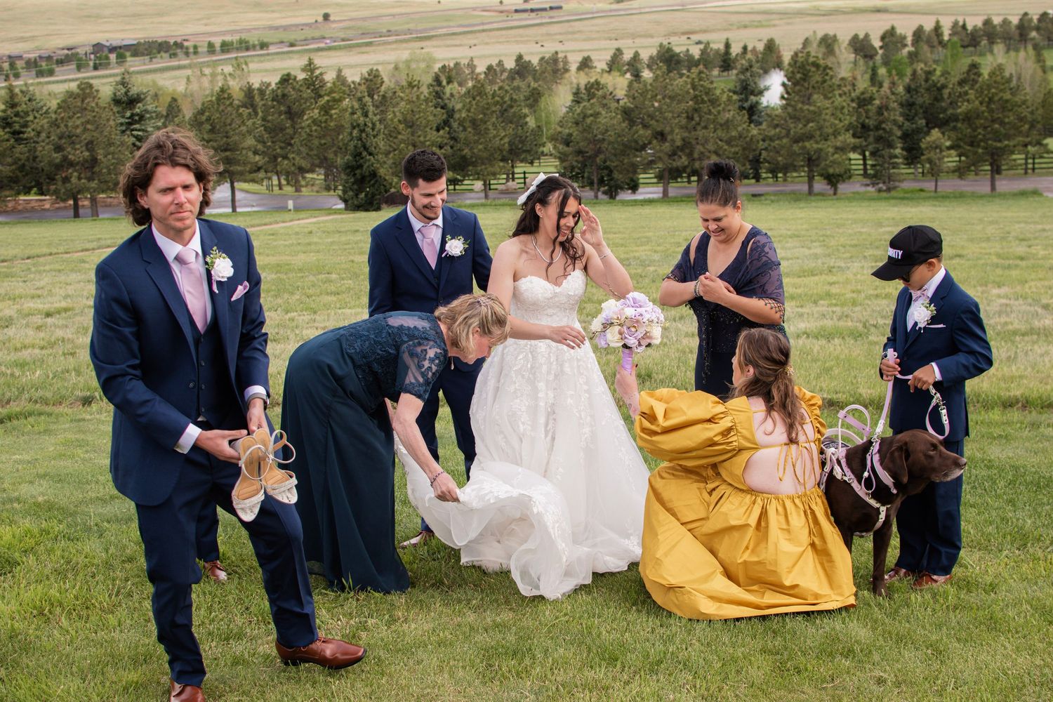 Wedding party helping bride with her gown on a green lawn with pine trees in background.