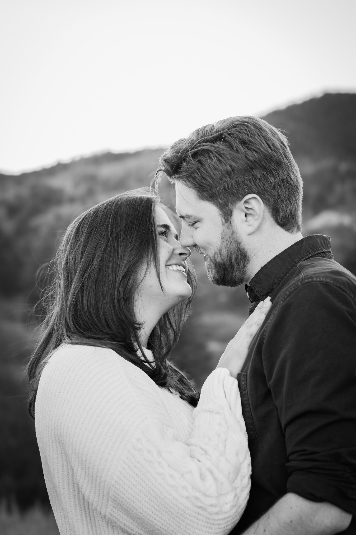 Black and white photo of a smiling couple sharing a romantic moment outdoors with rolling hills in the background.