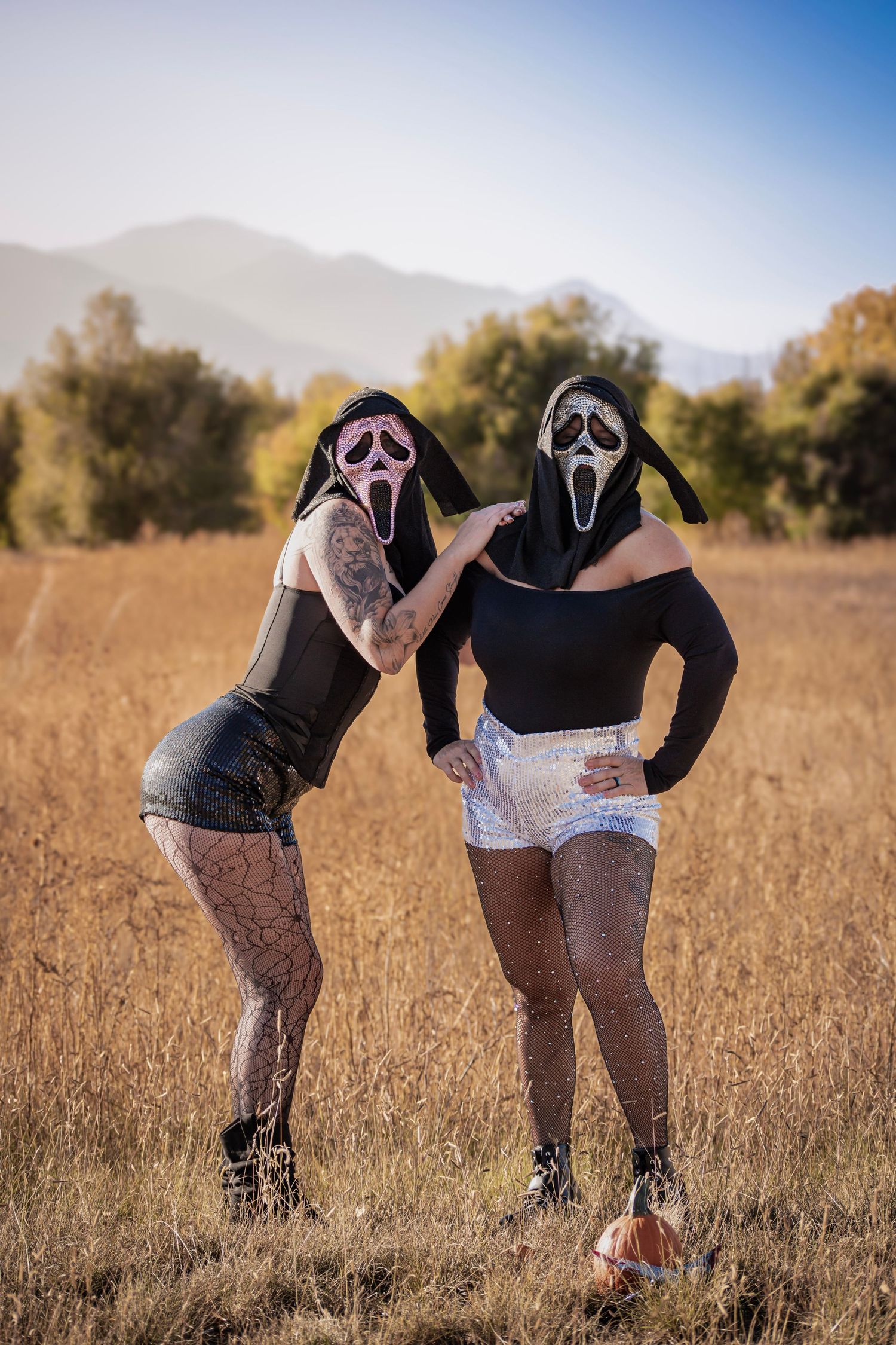 Two people wearing Ghostface Halloween masks and costumes posing in a dry field with mountains in background.