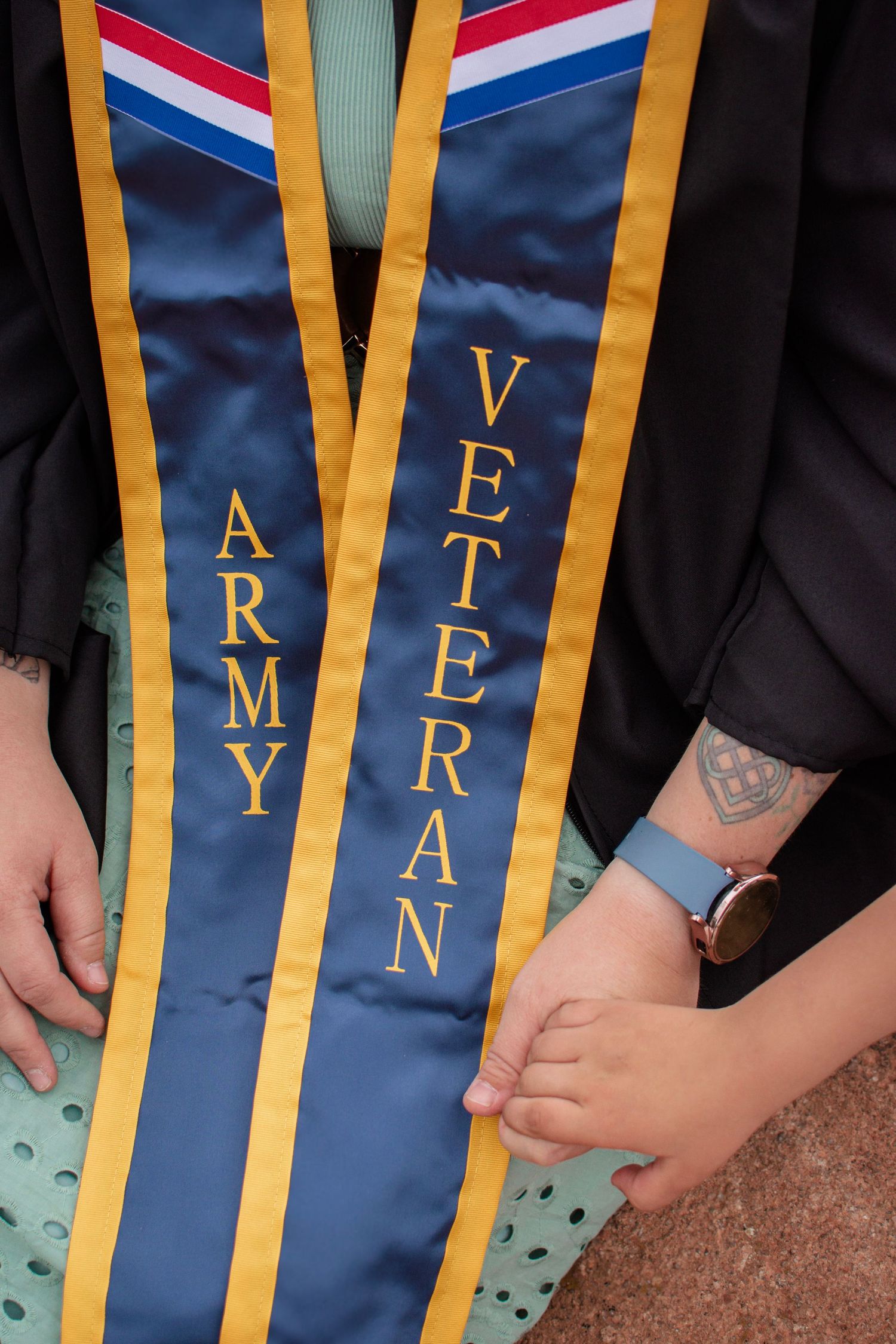 Army Veteran graduation stole in blue and gold worn over academic regalia, with a child's hand visible.