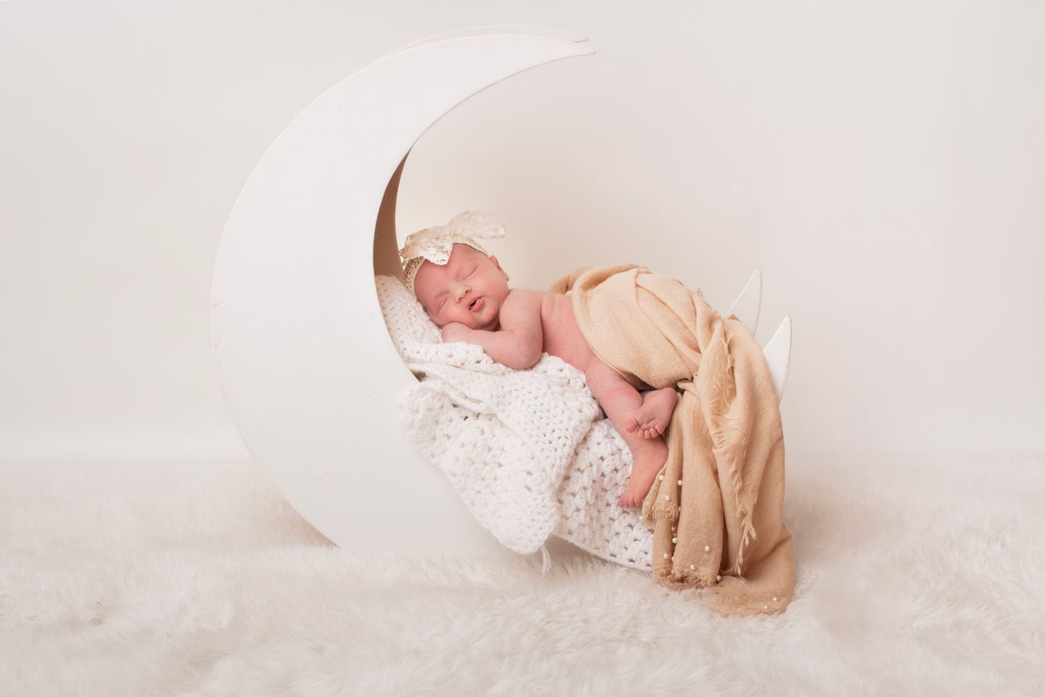 Newborn baby sleeping peacefully on a white crescent moon prop with soft blankets in a studio setting.