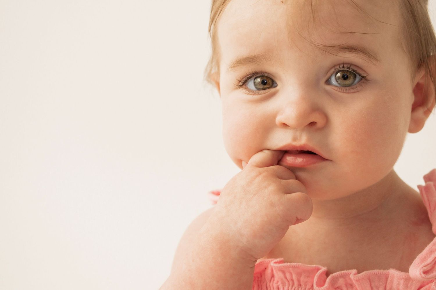 Cute baby girl in pink ruffled outfit with fingers in mouth, looking curious against white background at Luminous Creative Studios.