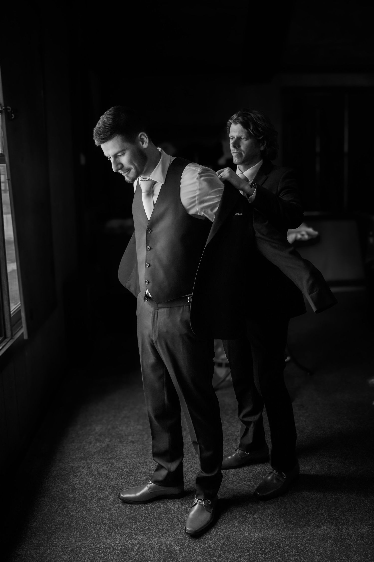 Black and white photo of groomsman helping groom put on suit jacket before wedding ceremony.