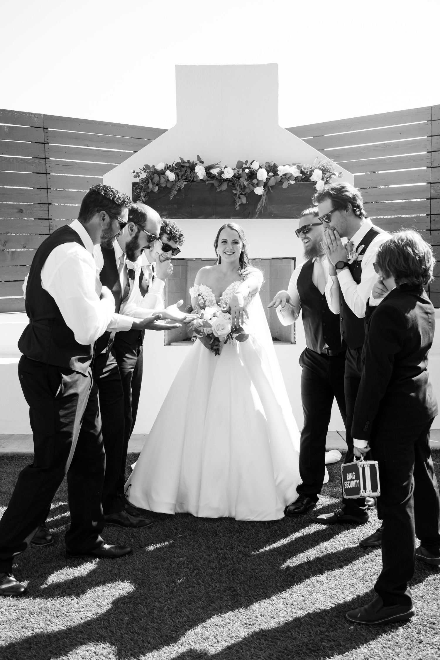 Black and white wedding photo of bride with bouquet and groomsmen in suits gathered around an outdoor altar.