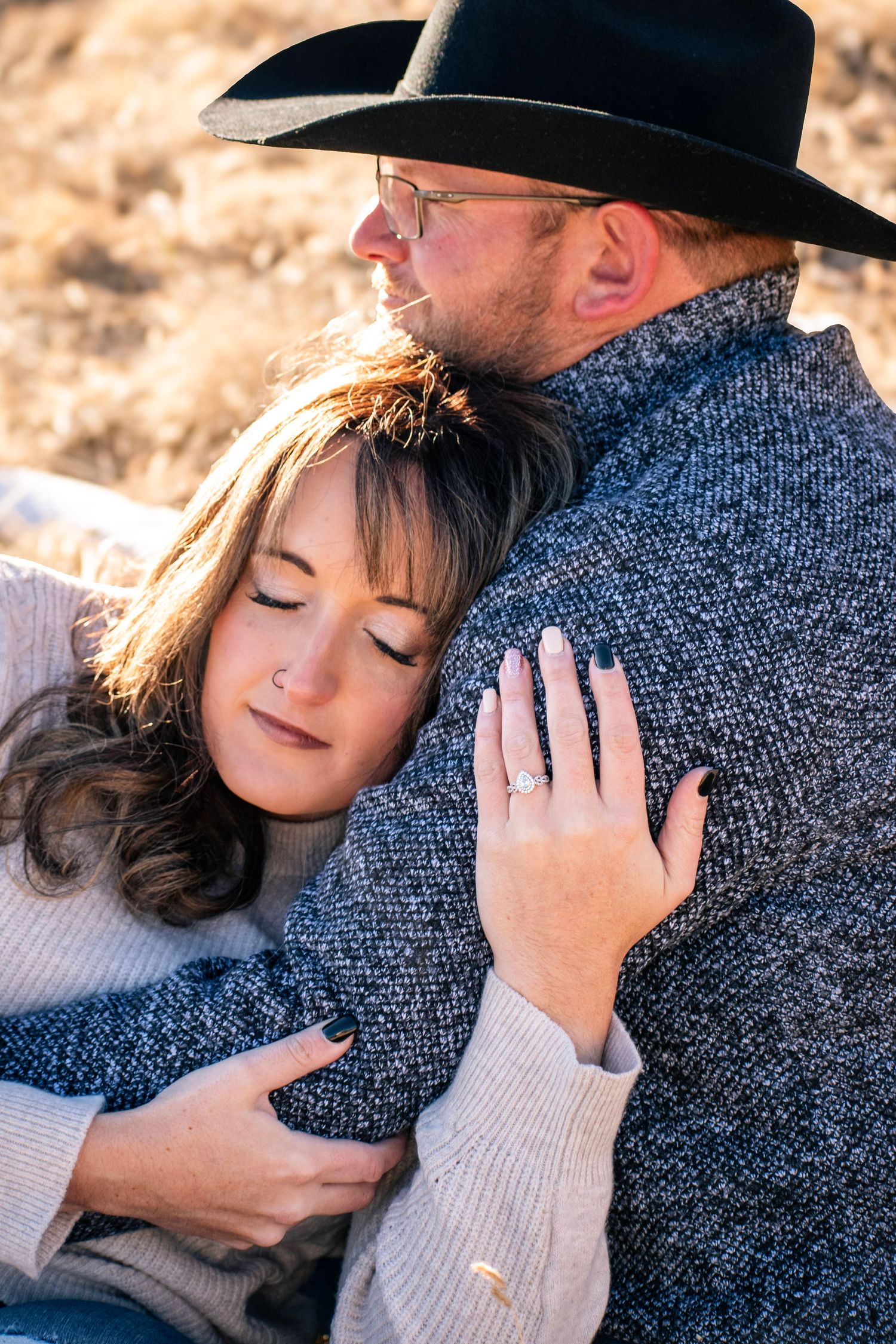 Couple embracing outdoors, woman with engagement ring resting head on man wearing cowboy hat in warm sunlight.