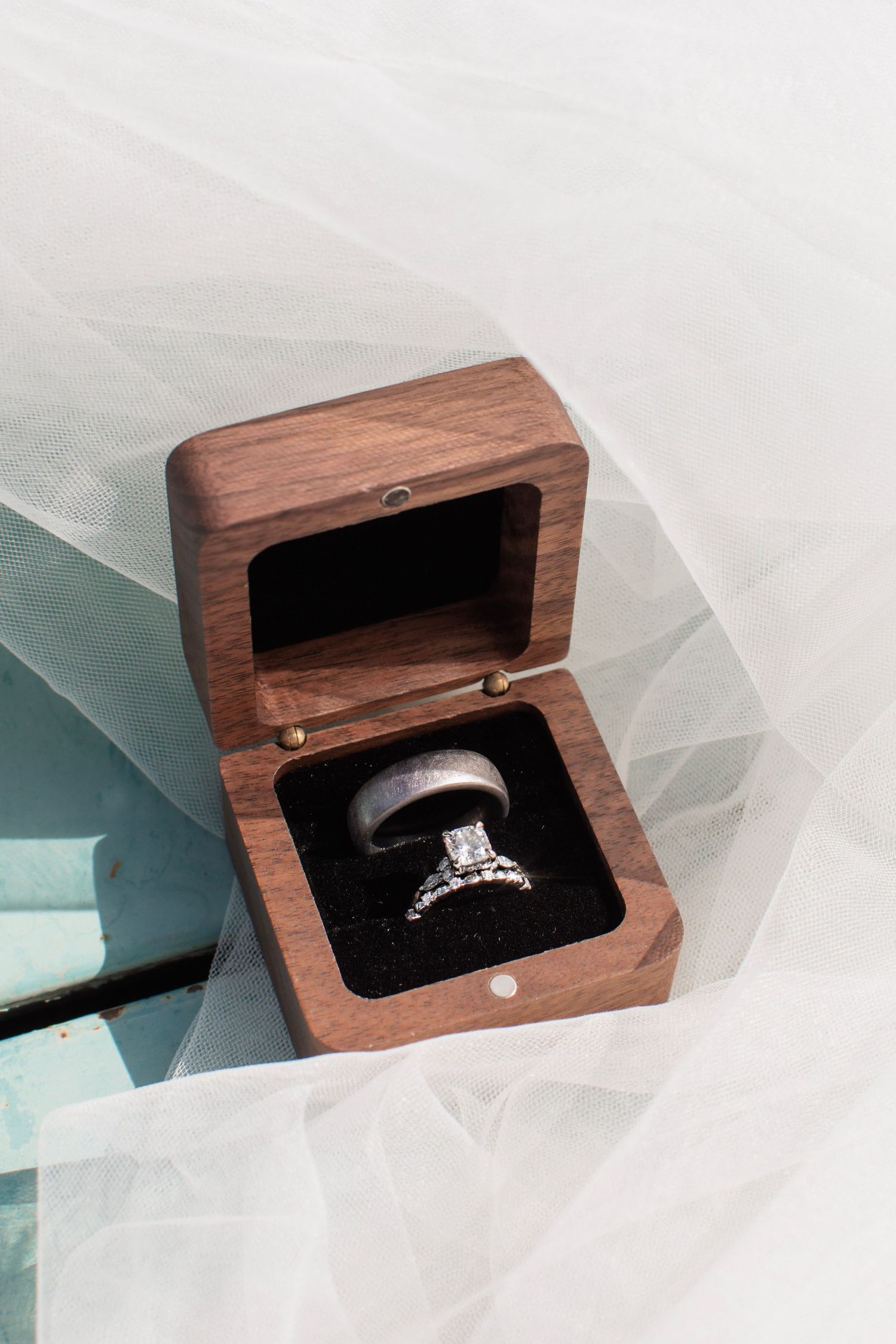 Diamond engagement ring and silver wedding band displayed in an open wooden ring box on a white bridal veil.