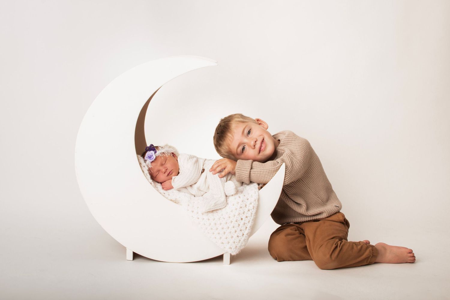 Smiling young boy in tan sweater rests his head on a white crescent moon prop holding a sleeping newborn baby in a purple flower headband.