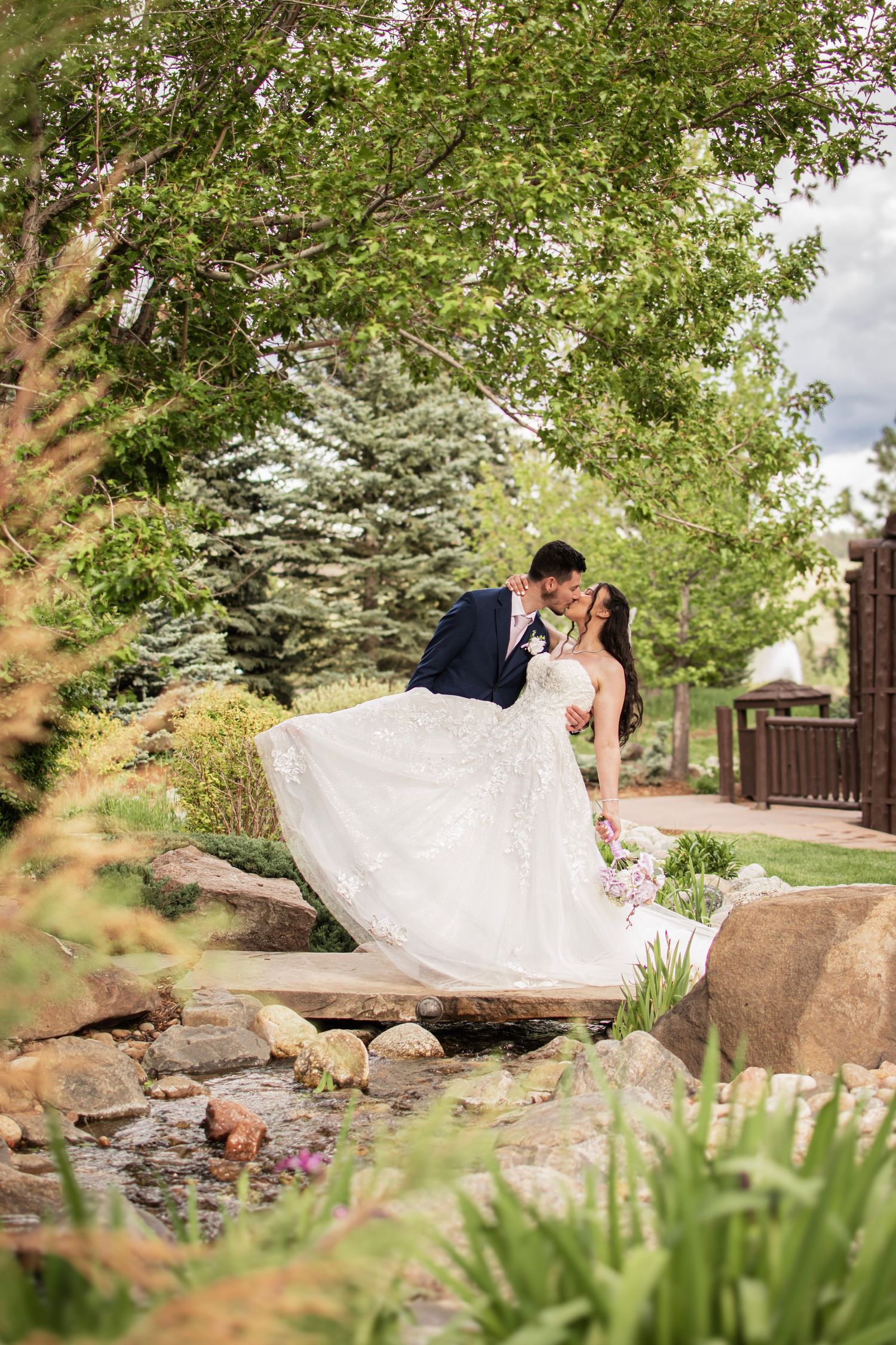 Bride and groom share a romantic kiss in a lush garden setting near a stone bridge on their wedding day.