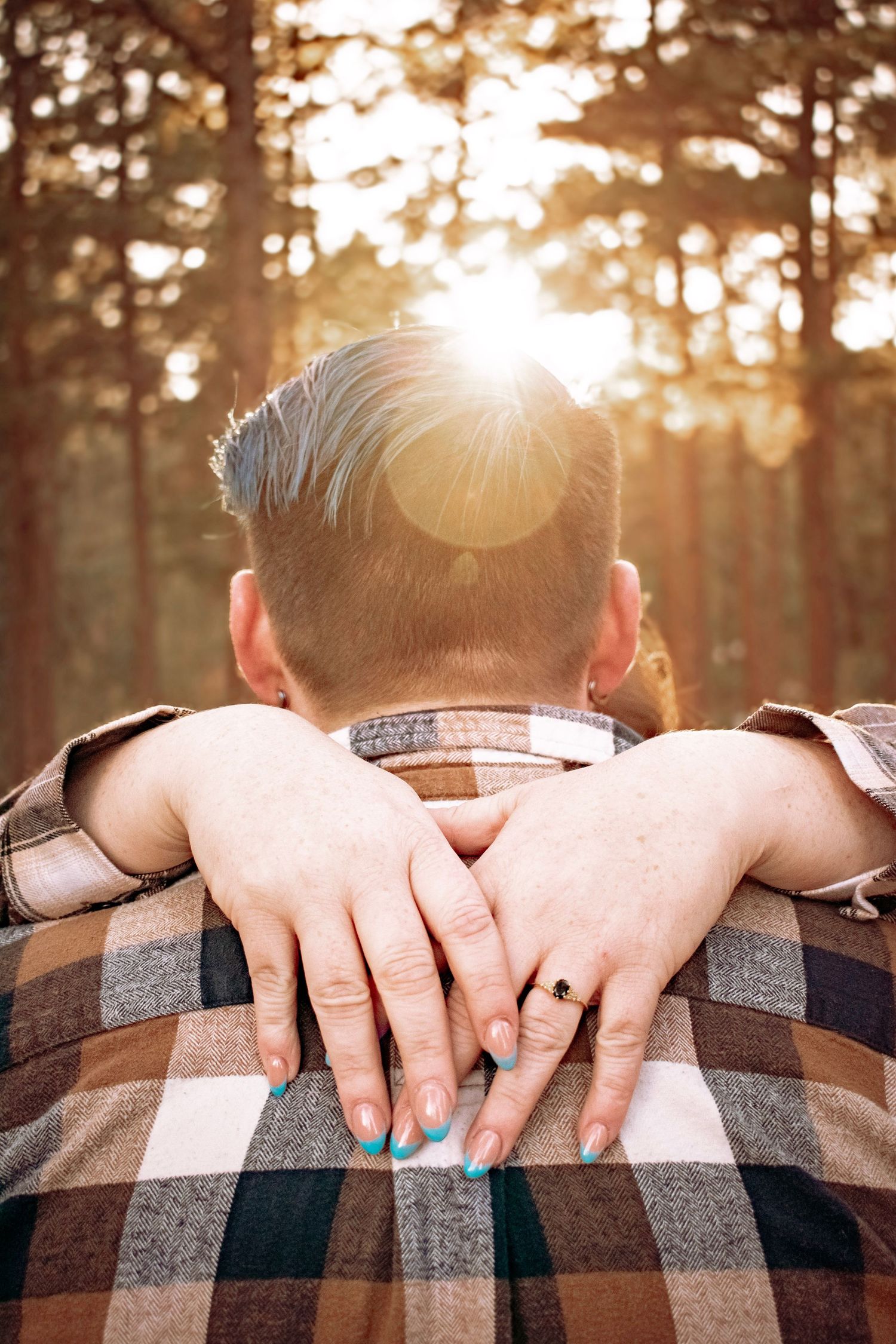 Couple embracing in a sunlit forest, woman's hands with teal nails and engagement ring resting on man's plaid shirt.