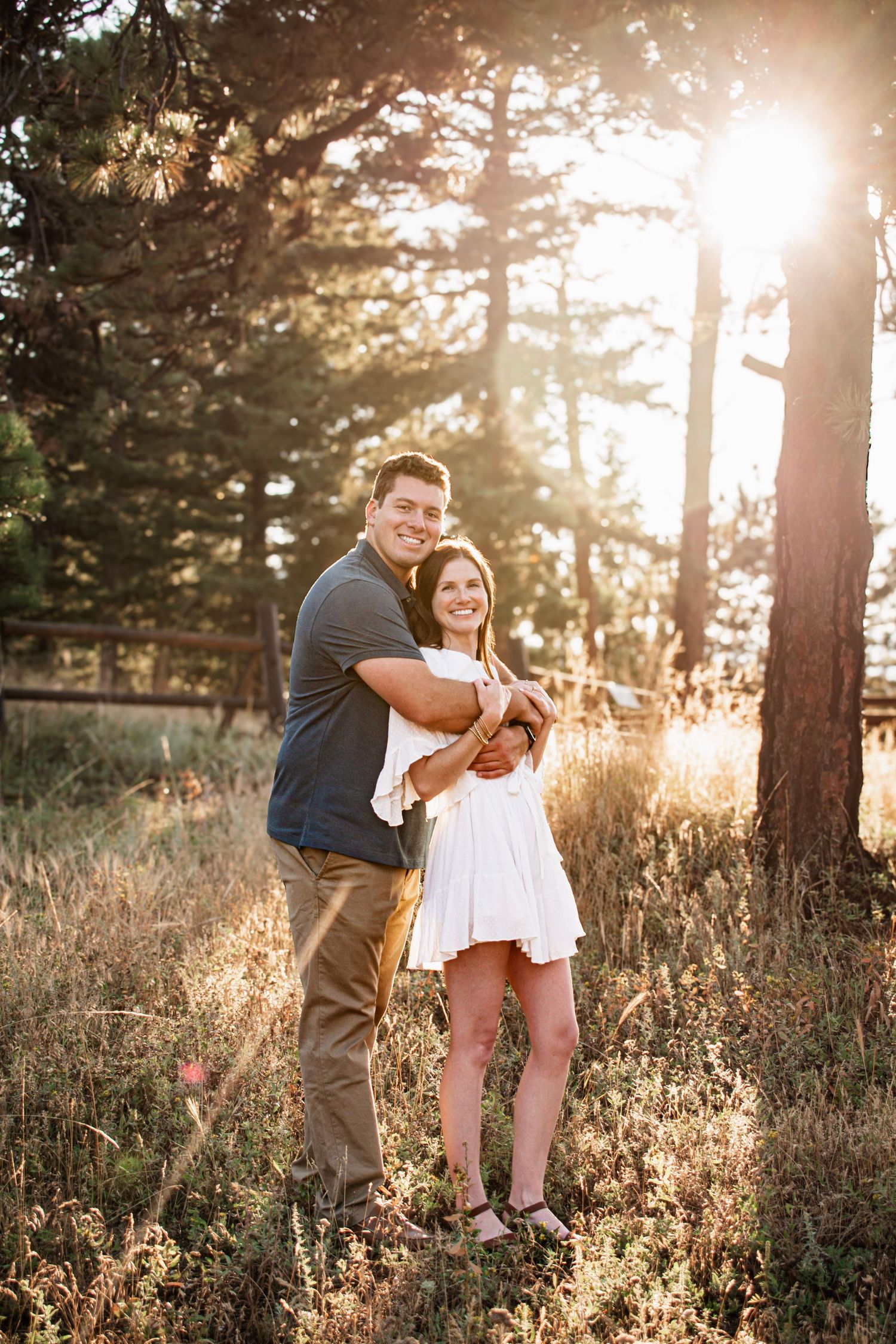 A couple embraces in a sunlit forest meadow, the woman in a white dress and man in a gray shirt, surrounded by tall pine trees.