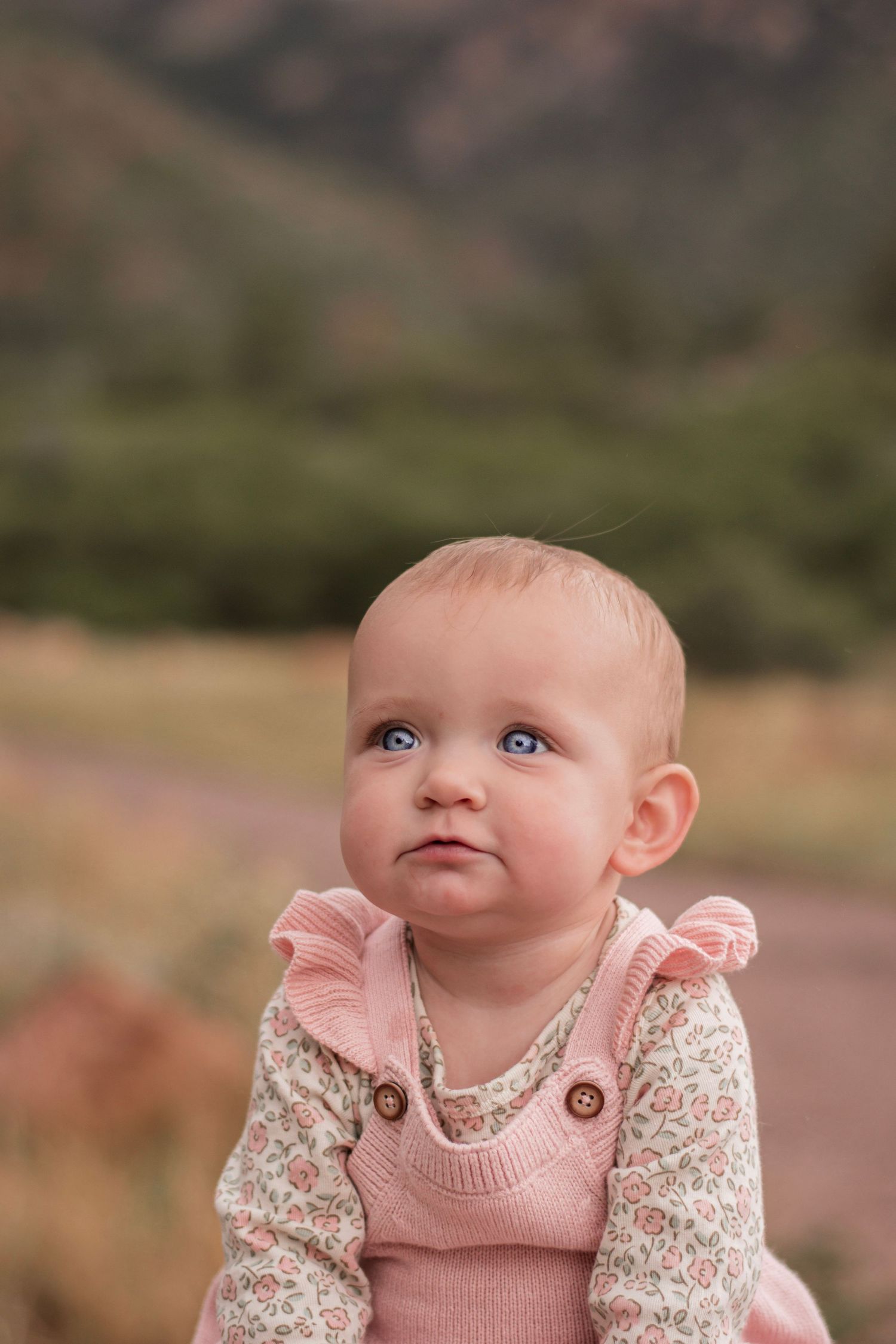 Blue-eyed baby girl in pink knit overalls and floral top sitting outdoors with mountain landscape background.