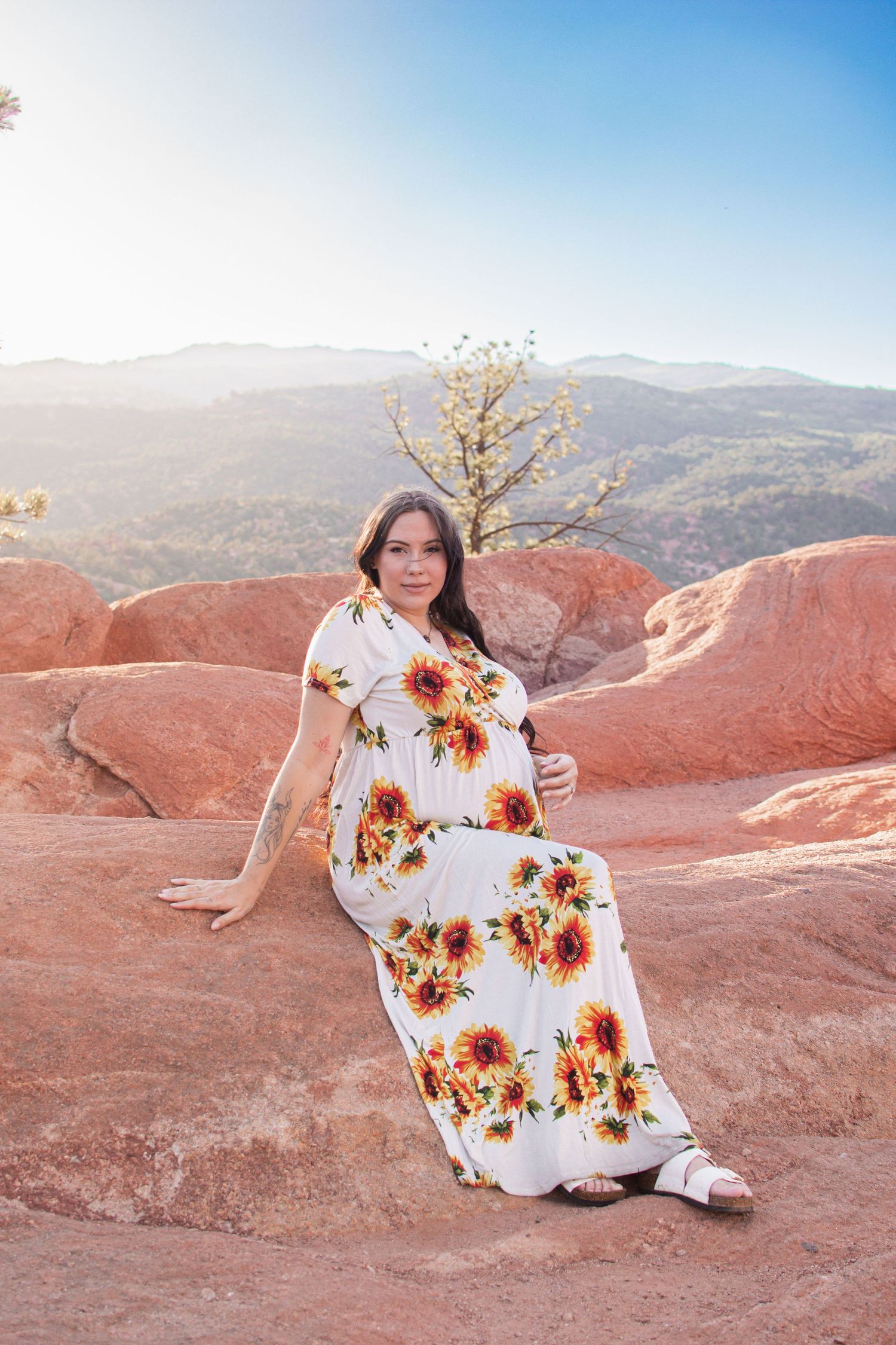 Woman in a sunflower print maxi dress sits on red sandstone rock with hazy mountains and a lone tree in the background at sunset.