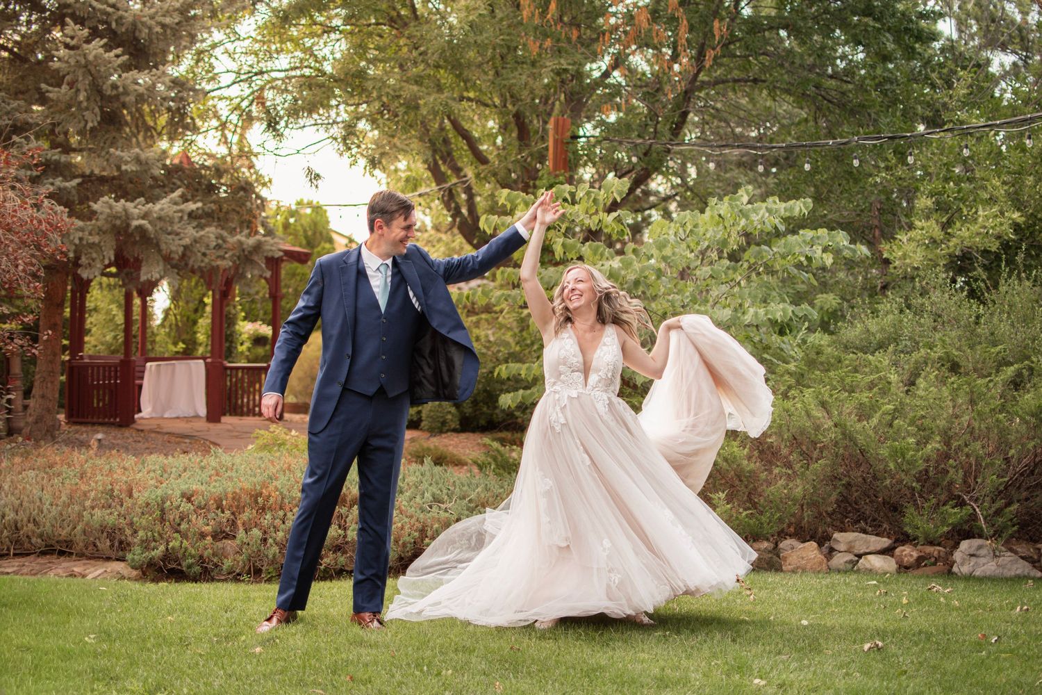 A joyful bride and groom dance outdoors on lush green grass, surrounded by trees, as she twirls in her flowing white gown.