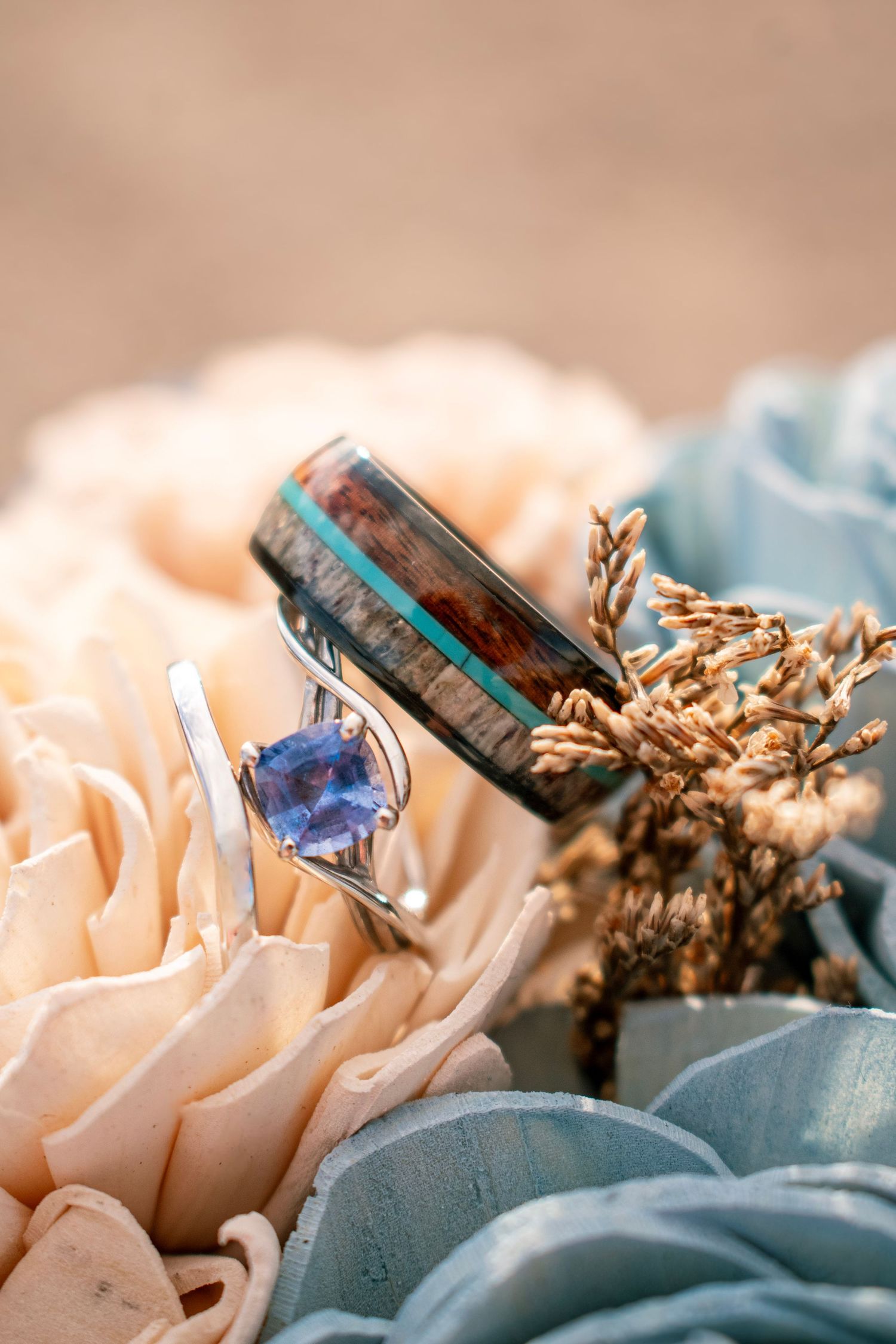 Two wedding rings rest on pastel flowers, featuring a blue gemstone engagement ring and a wood inlay band with turquoise accents.