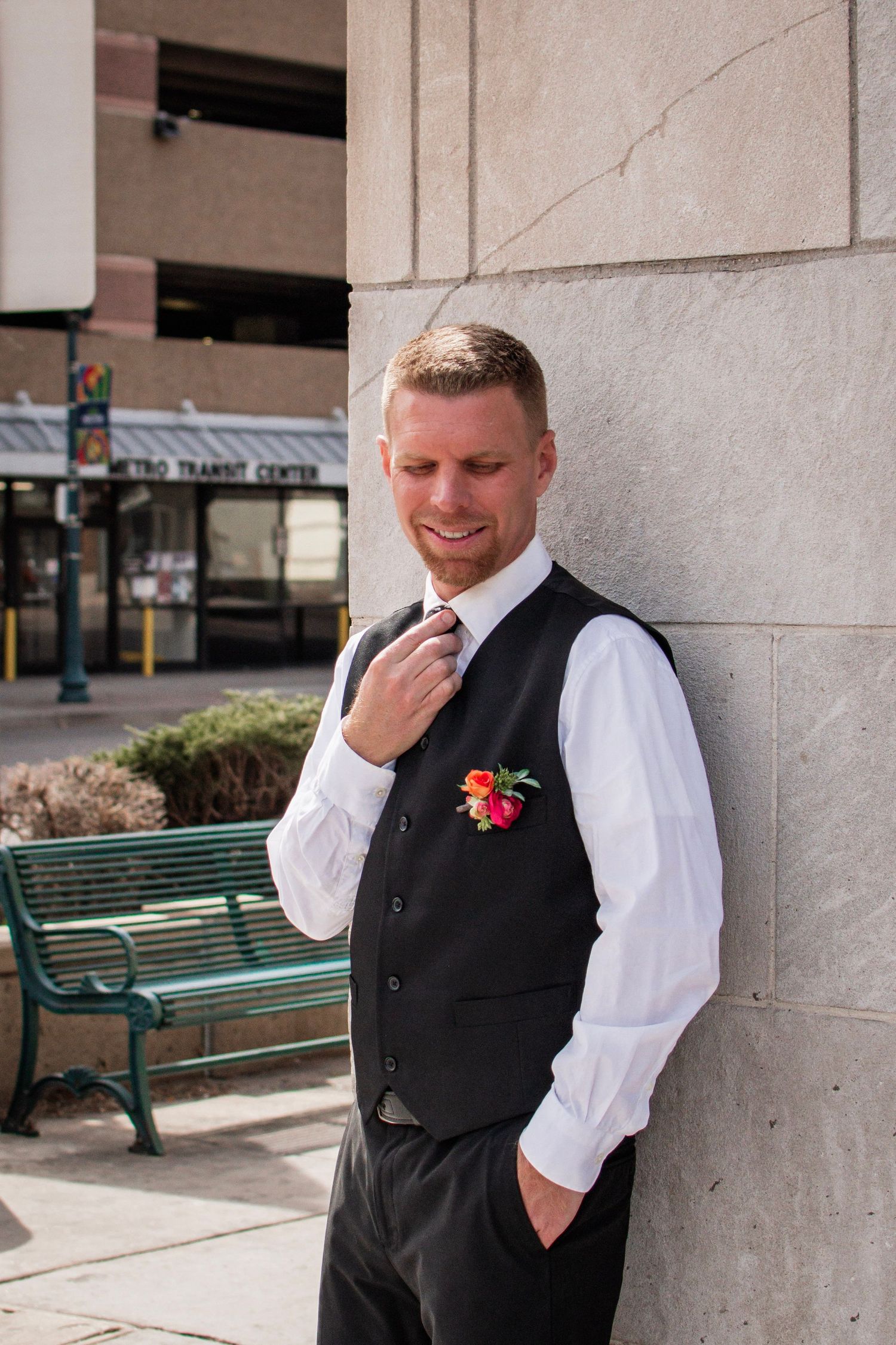 Smiling groom in black vest, white shirt, and floral boutonniere leans against stone wall outdoors near Metro Transit Center.
