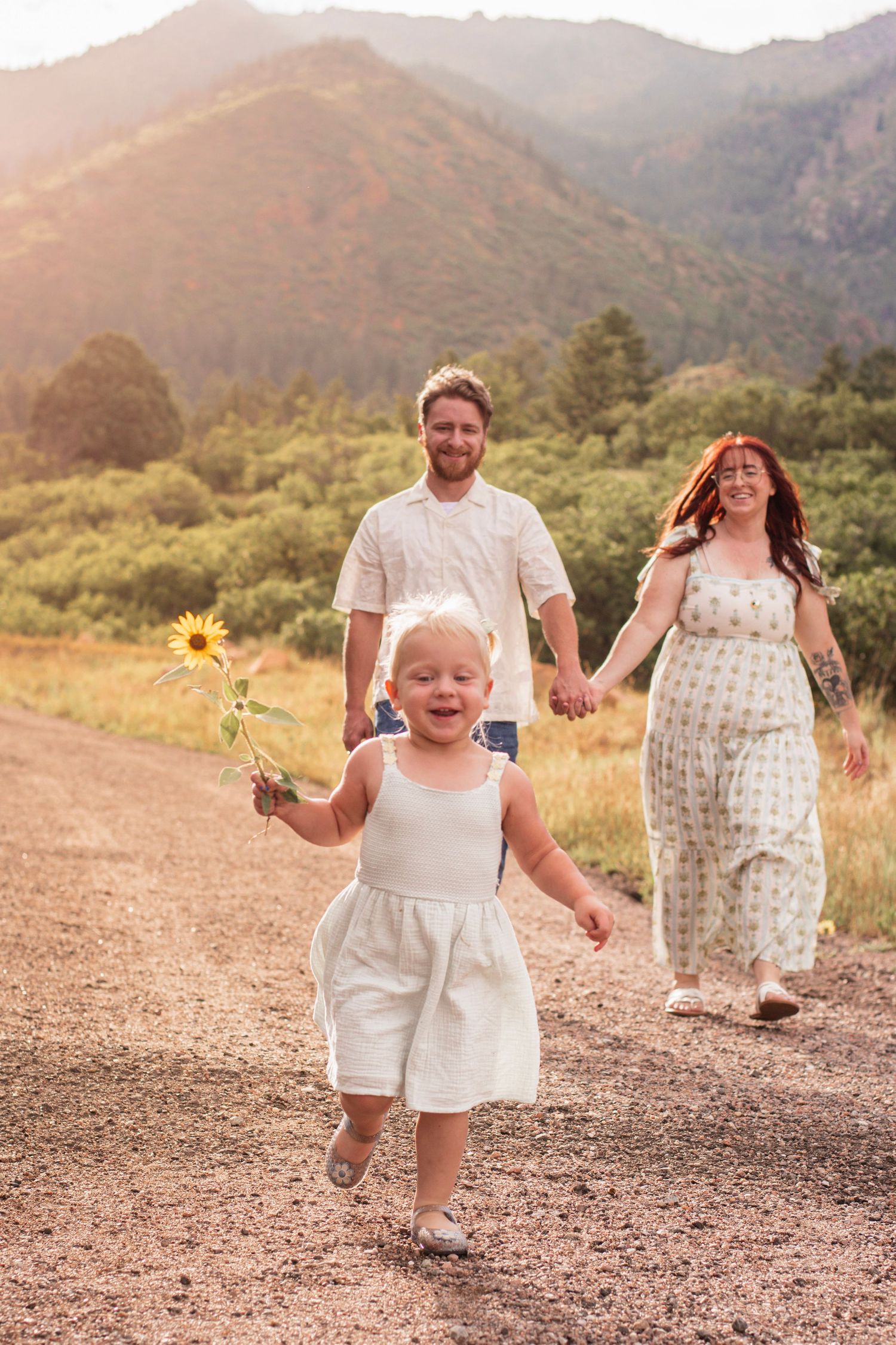 A joyful toddler runs with a sunflower as her pregnant mother and father walk hand-in-hand on a scenic mountain path.