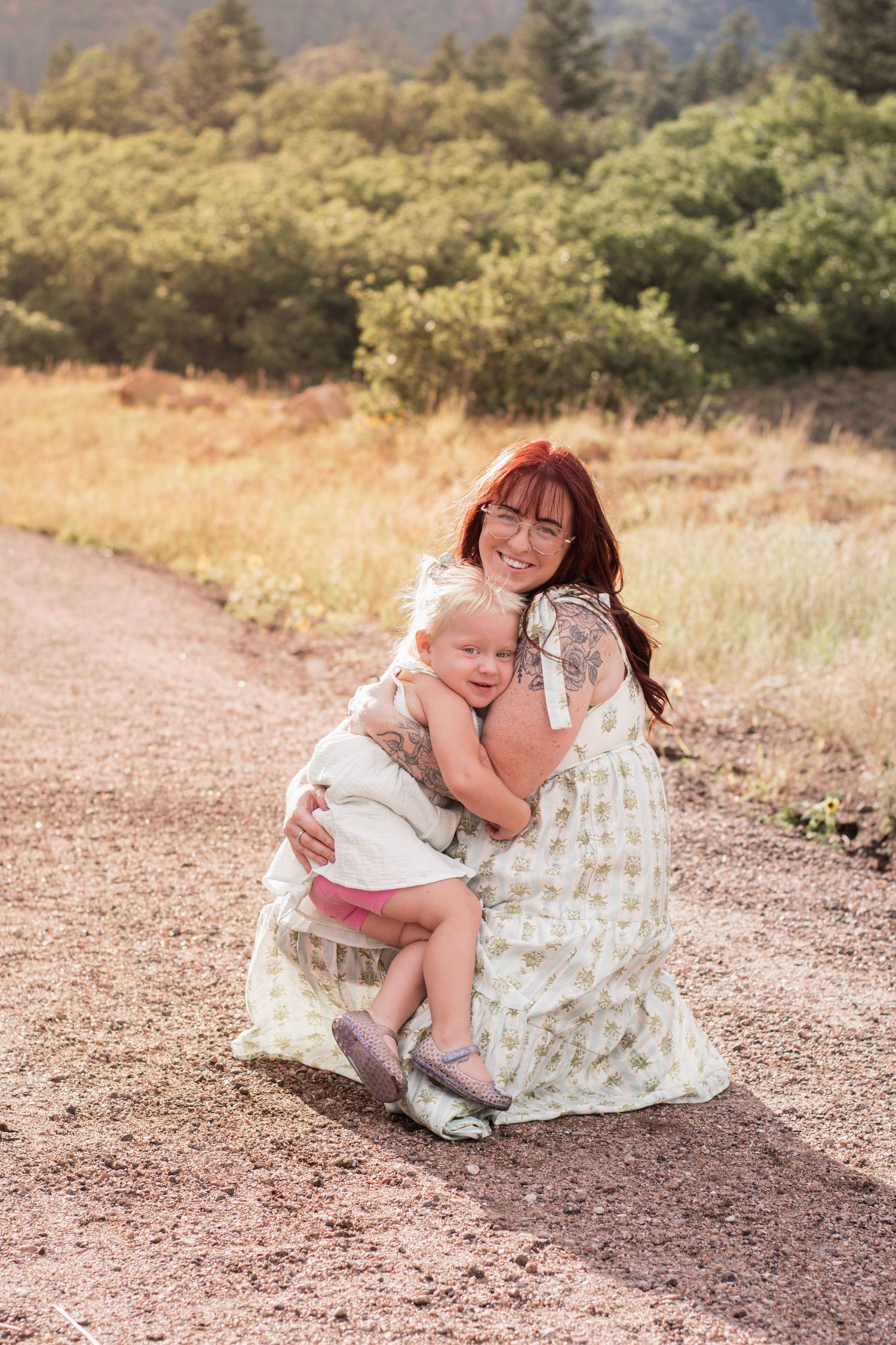 A woman with red hair crouches on a dirt path hugging a toddler girl, surrounded by green trees.