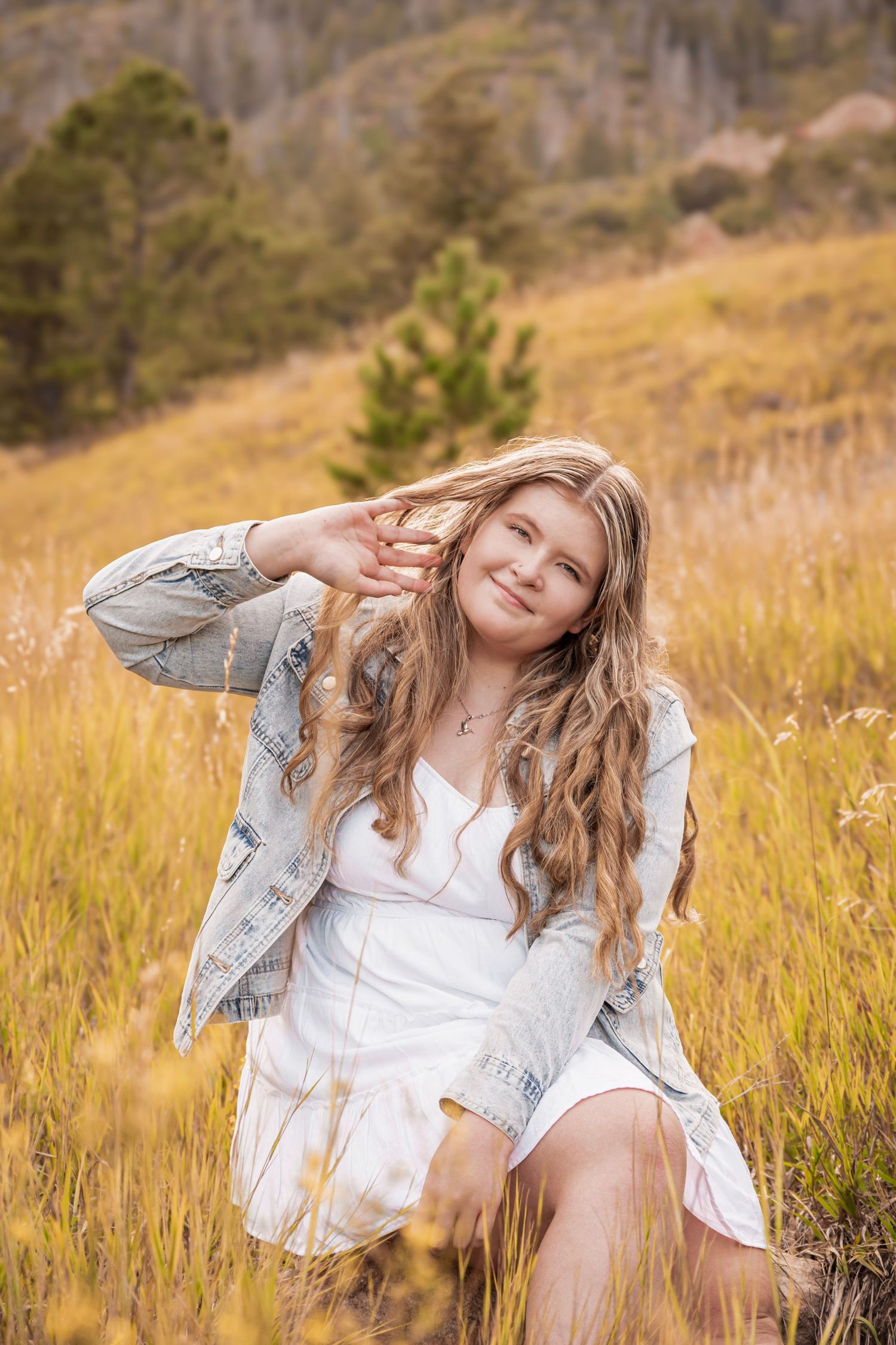 Young woman with long wavy hair sits in golden meadow wearing white dress and denim jacket, smiling outdoors.