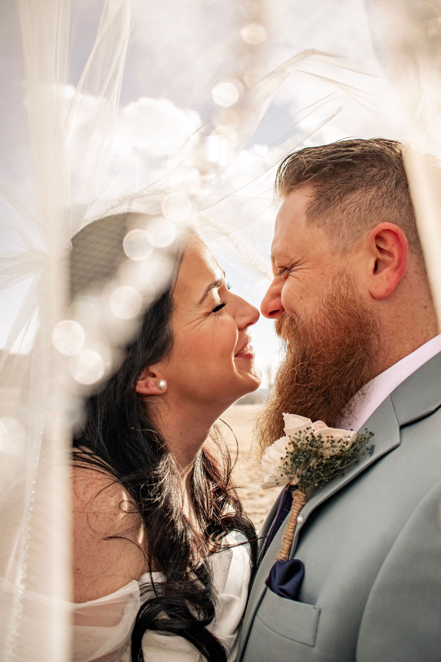 Bride and groom sharing a romantic moment under a veil, smiling at each other on their wedding day.