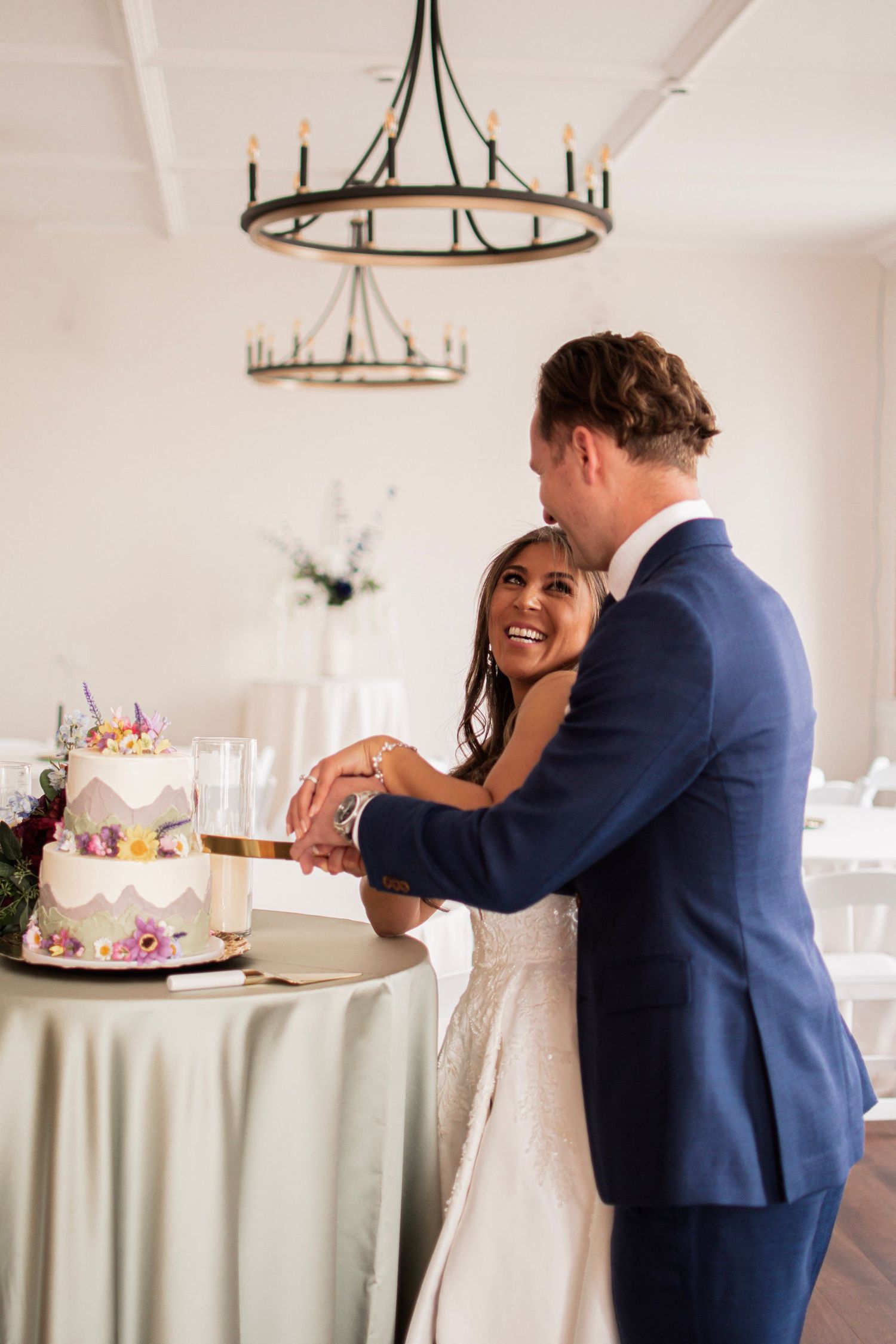 Bride and groom cutting their floral wedding cake together in an elegant white reception venue with chandelier.
