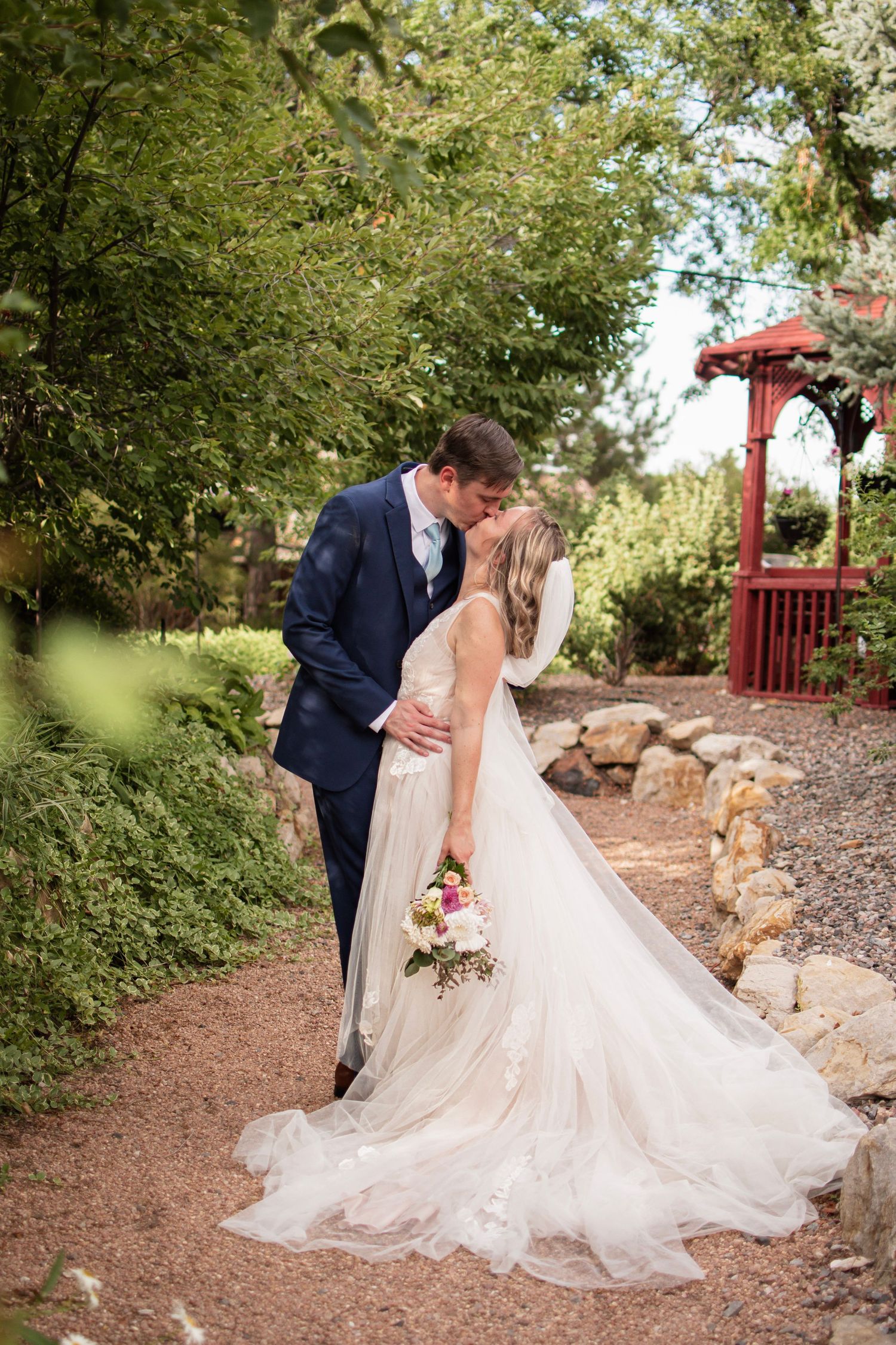 Bride and groom share a romantic kiss in a garden, bride in flowing white gown holding colorful bouquet, groom in navy suit.