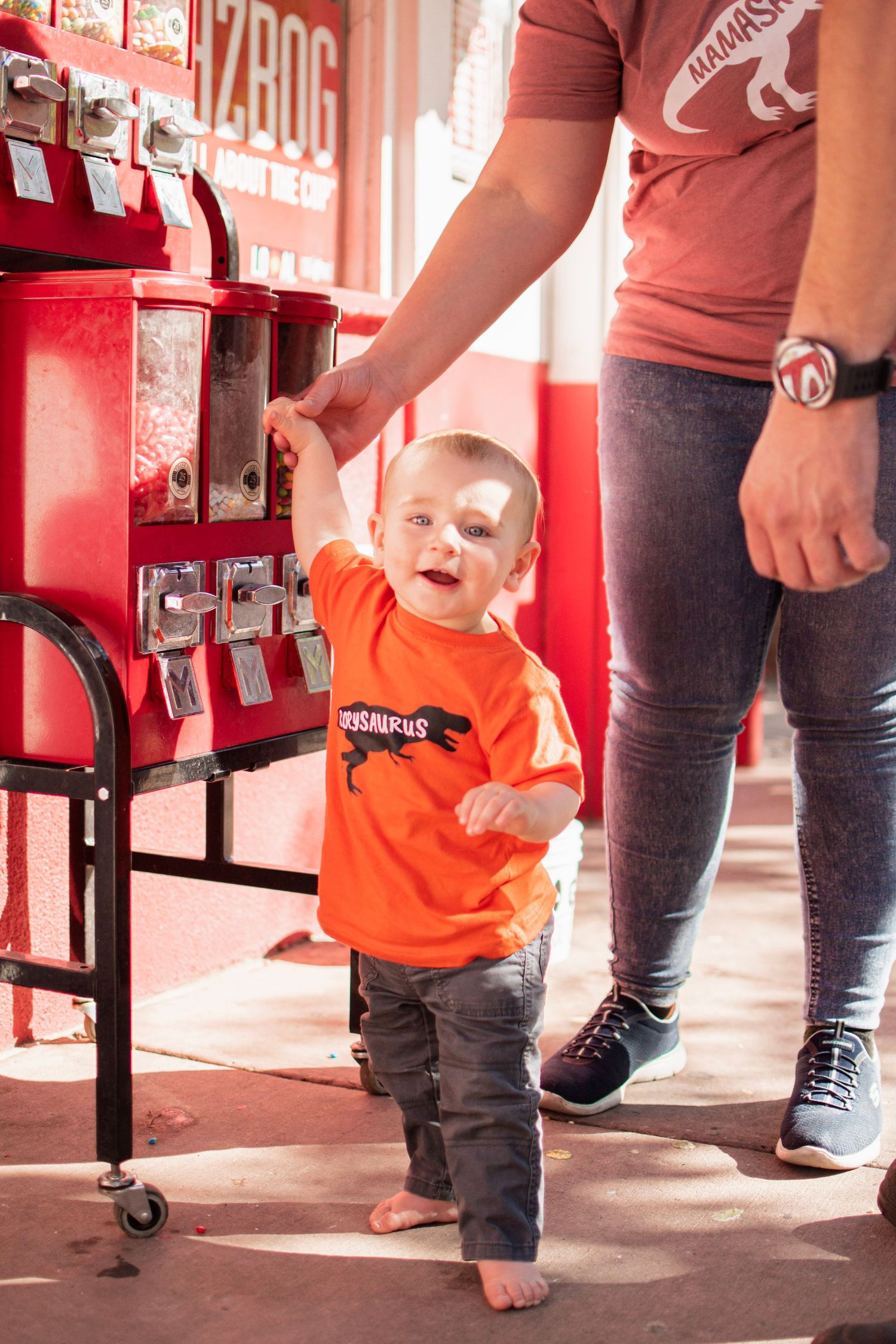 A toddler in an orange dinosaur shirt holds an adult's hand near red gumball machines at the Penny Arcade in Manitou Springs, CO.
