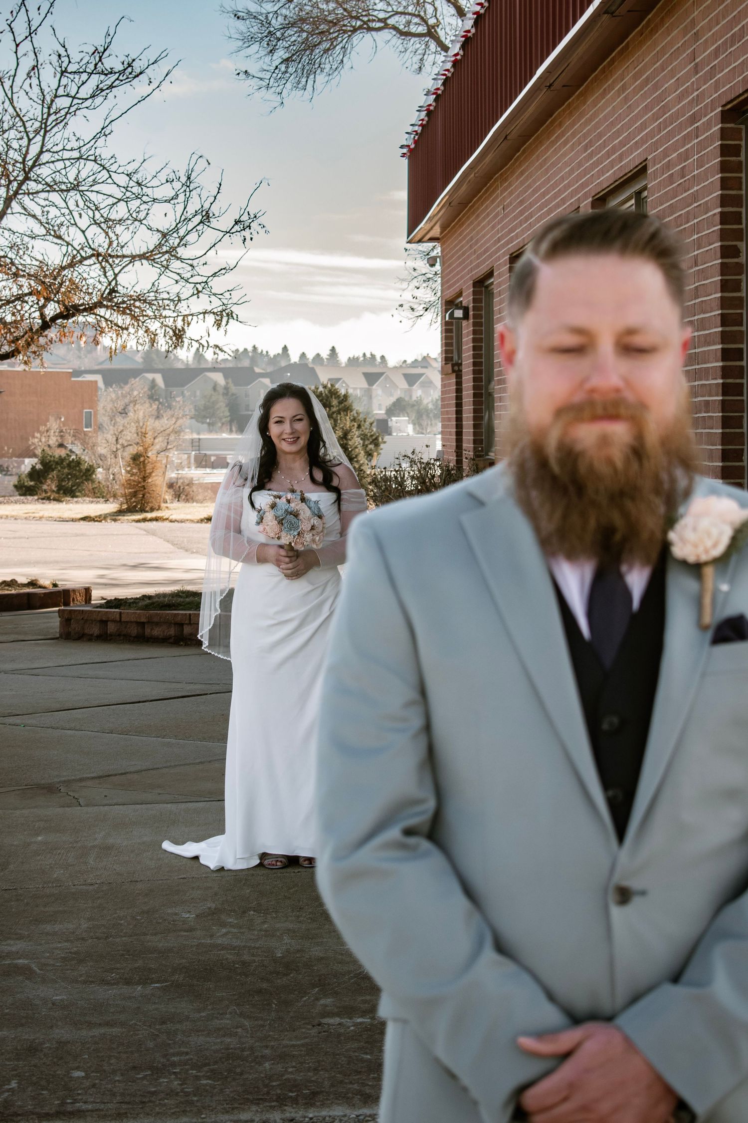 Bride in white gown smiles at bearded groom in light gray suit during outdoor first look at wedding venue.