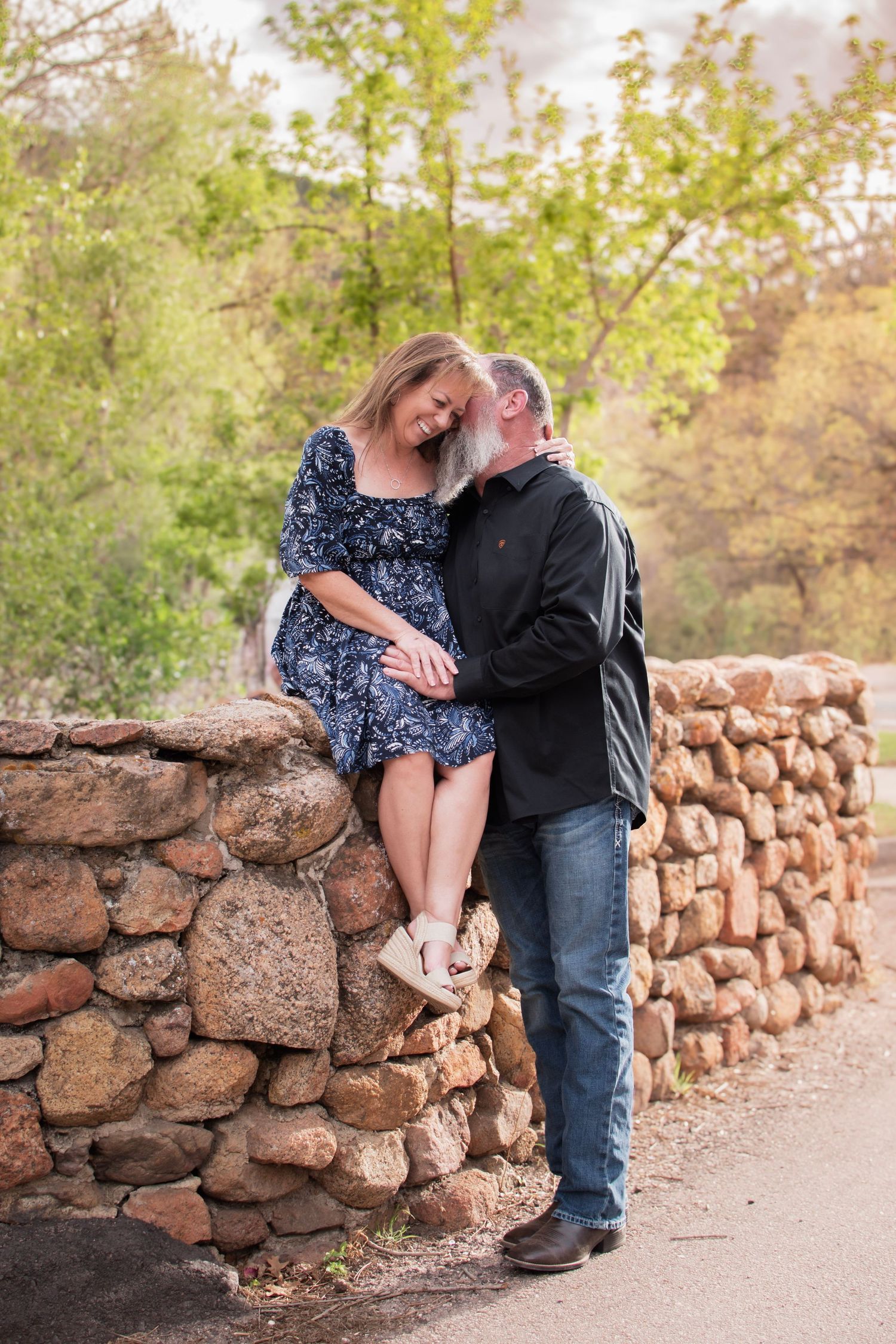 Couple sharing a tender moment on a rustic stone wall surrounded by lush green trees in an outdoor park setting.