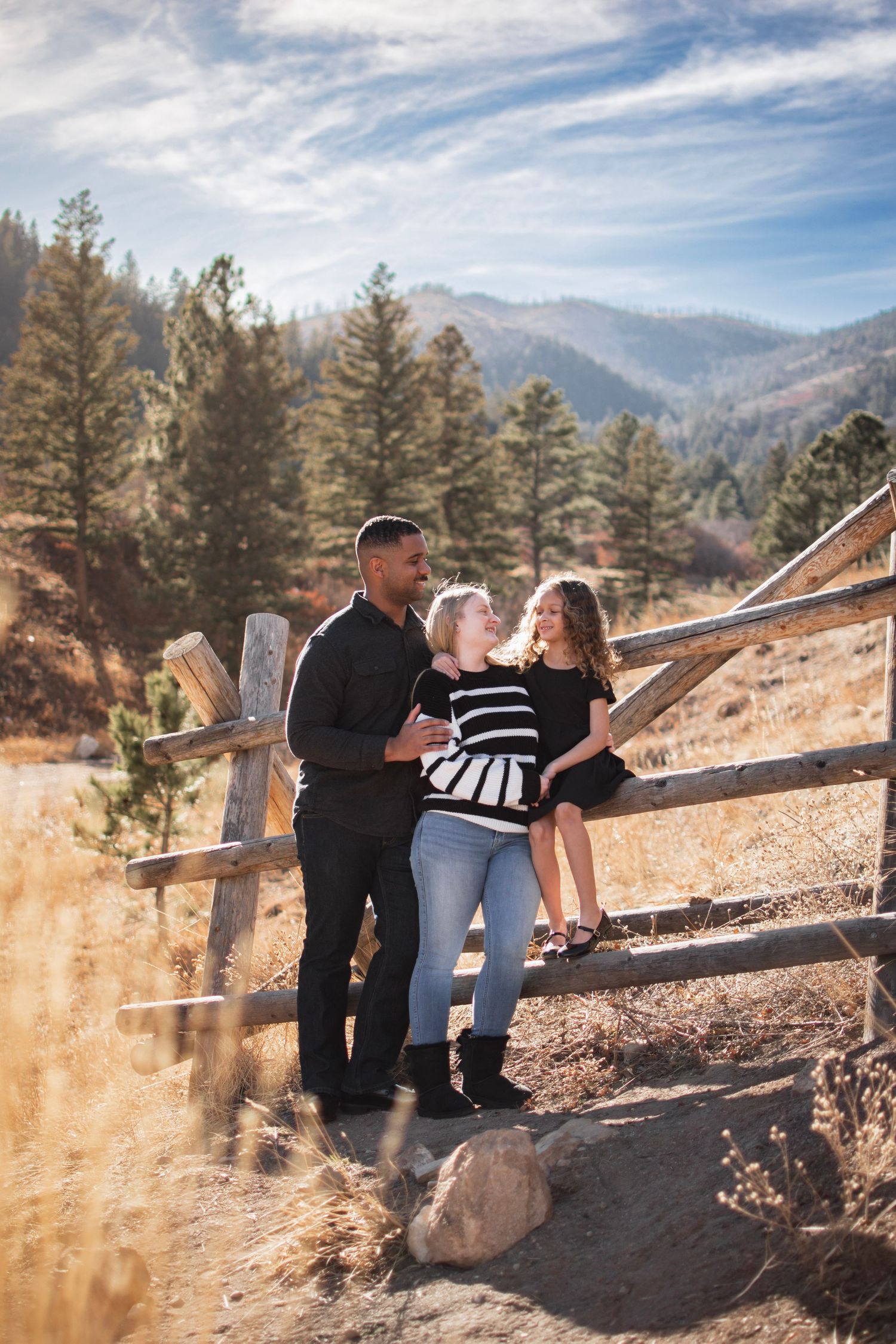 Family of three posing by rustic wooden fence in mountain landscape with pine trees and blue sky.