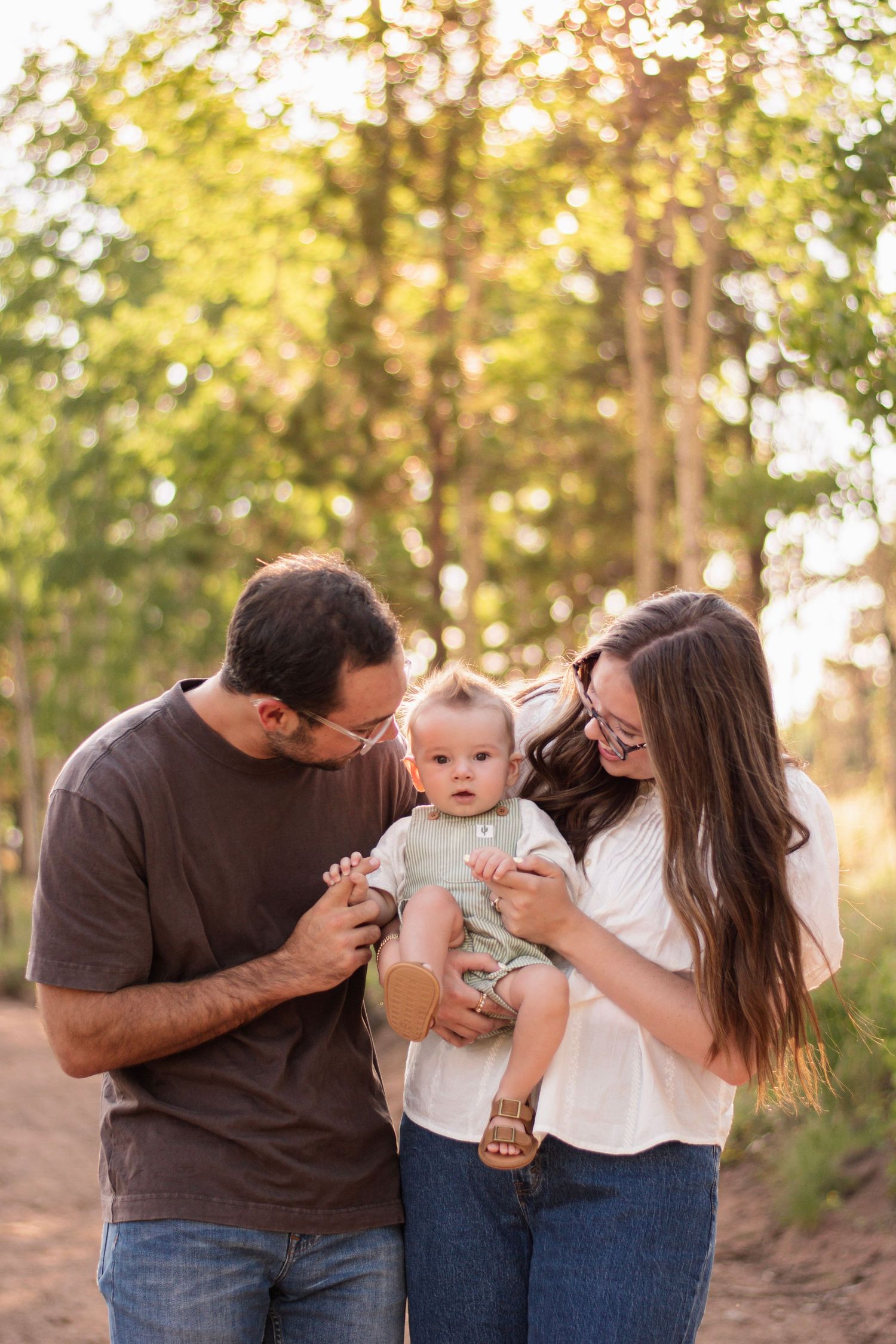 A young family poses outdoors on a sunny path, parents smiling at their baby in a green outfit, surrounded by trees.