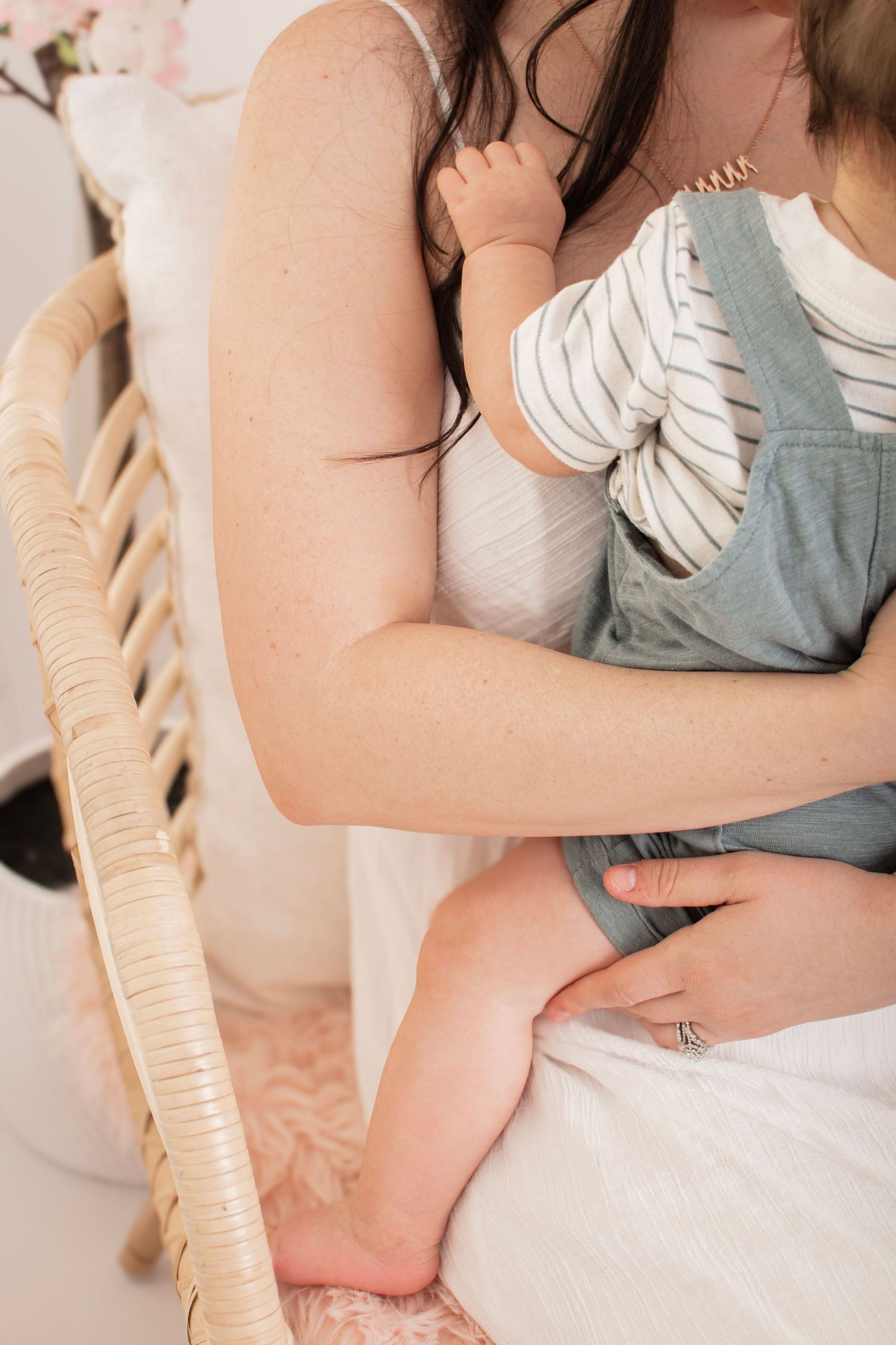 Mother holding baby in rocking chair, infant wearing striped shirt and green overalls, soft natural light nursery setting.