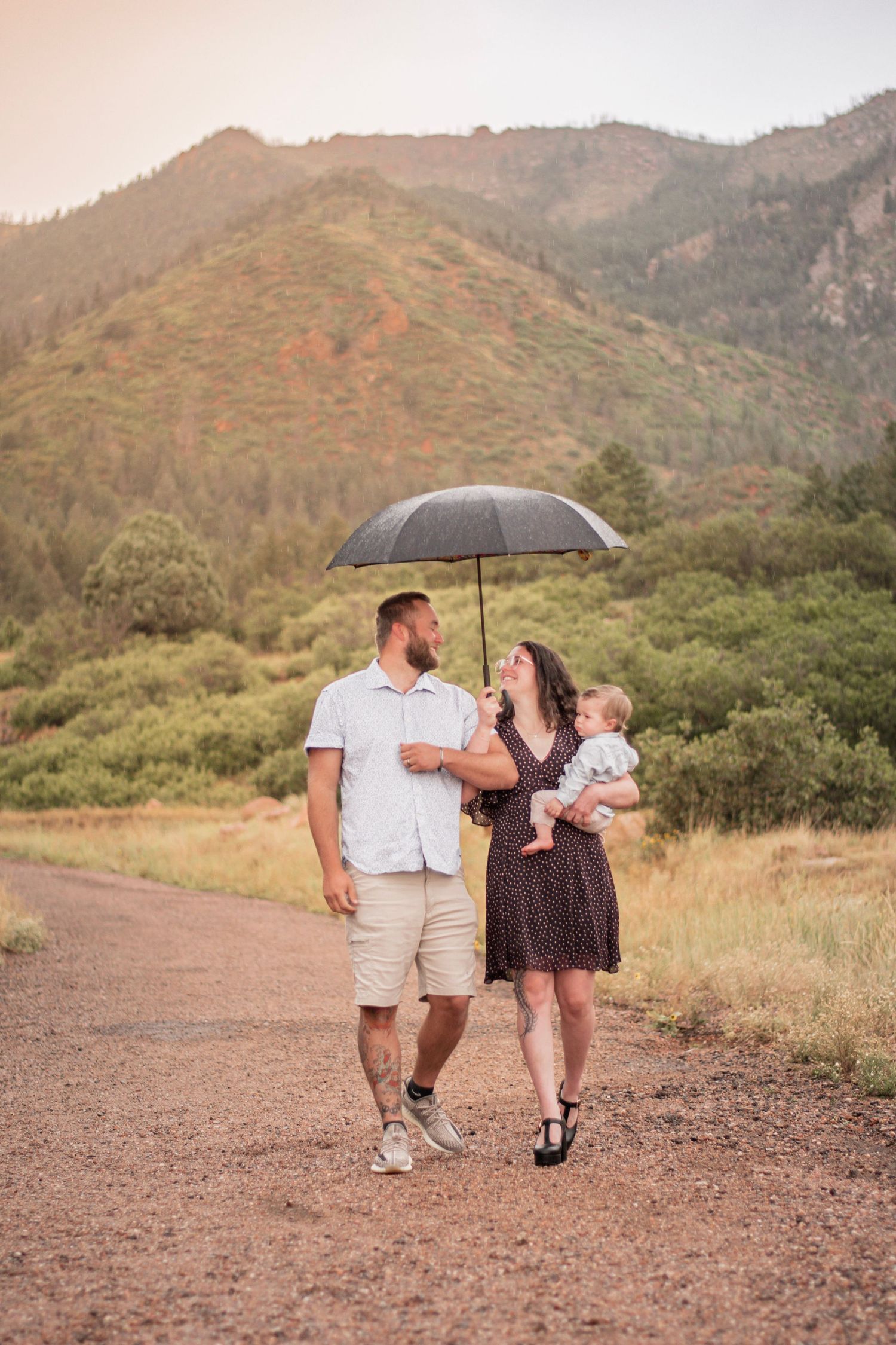 A couple walks with their baby under a black umbrella on a mountain trail surrounded by lush greenery At Blodgett Open Space.
