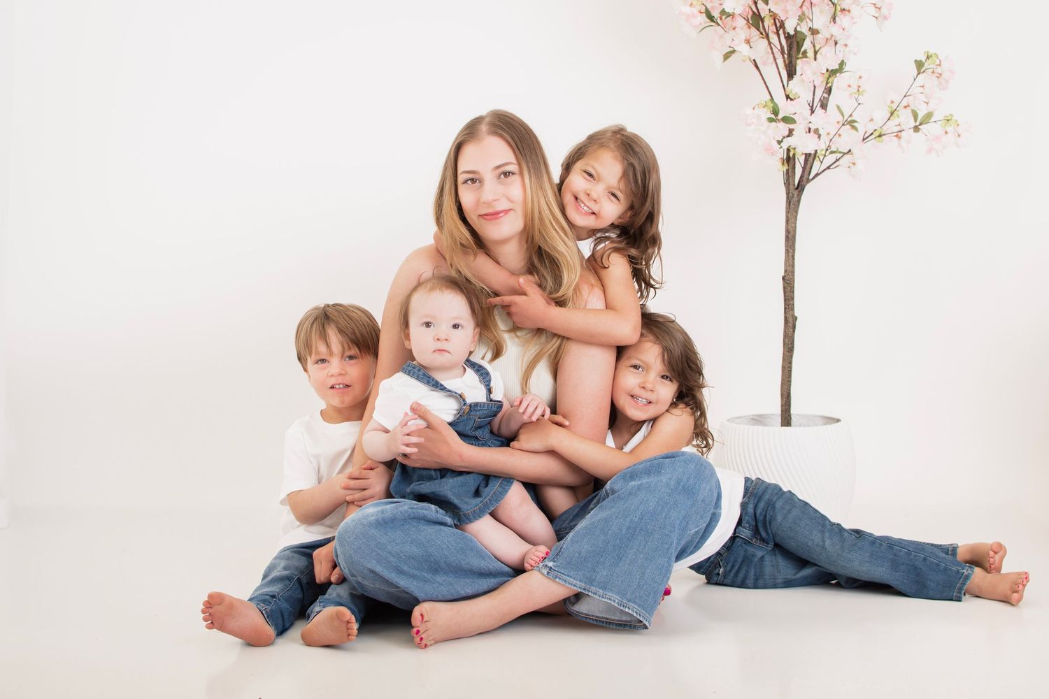 A woman sits on a white floor surrounded by four young children, all wearing jeans, near a blossoming tree.