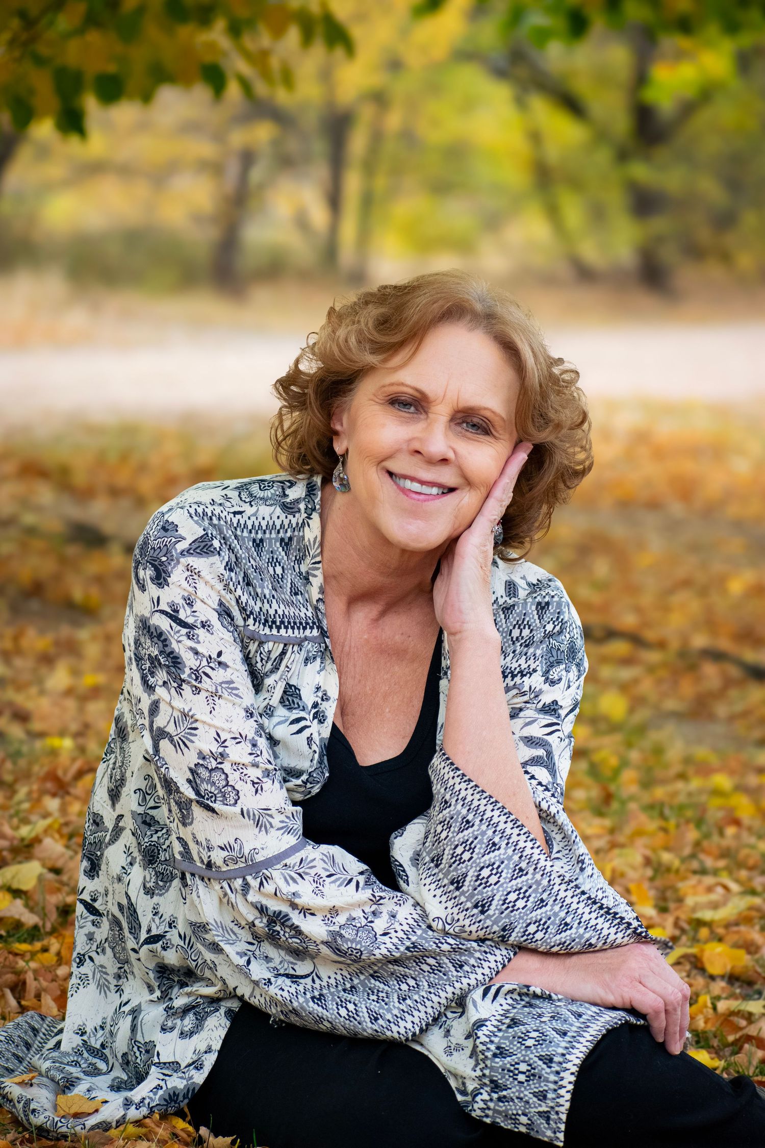Smiling middle-aged woman with curly hair sitting outdoors among autumn leaves wearing a floral kimono jacket.