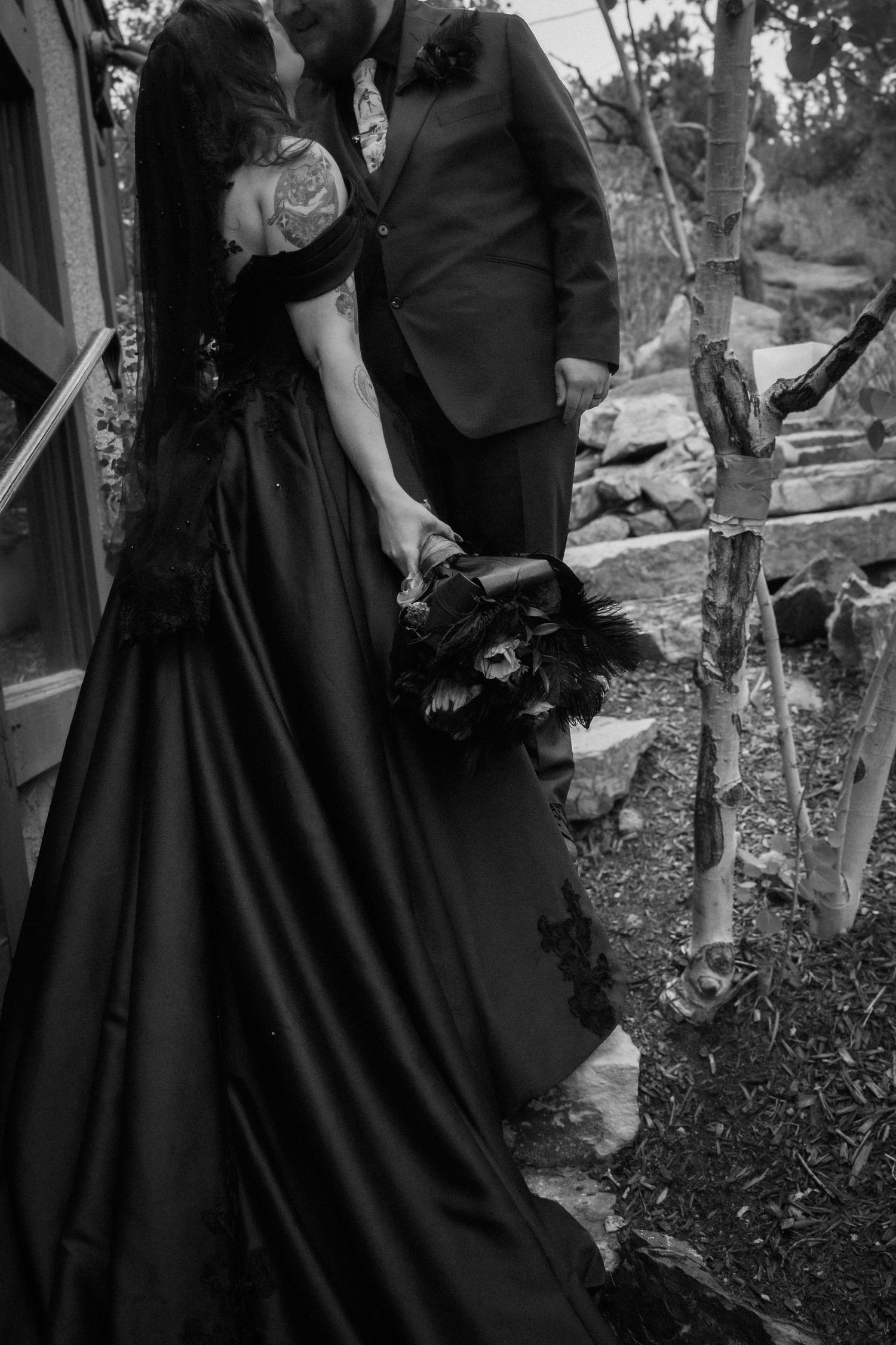 Black and white photo of a gothic wedding couple, bride in dramatic black gown holding dark bouquet outdoors.