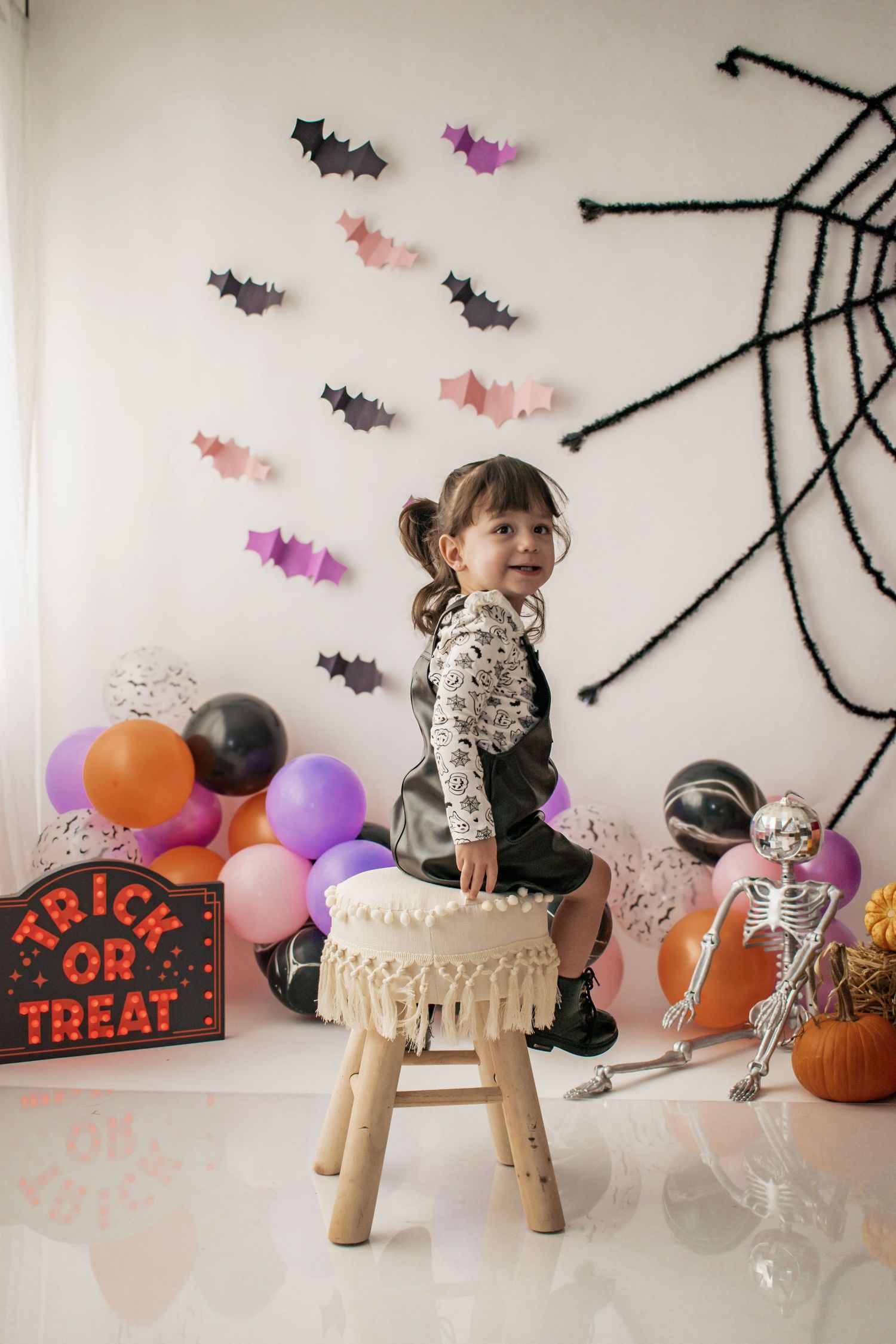 A young girl sits on a stool amid Halloween decorations including bats, a spiderweb, balloons, skeleton, and a Trick or Treat sign.