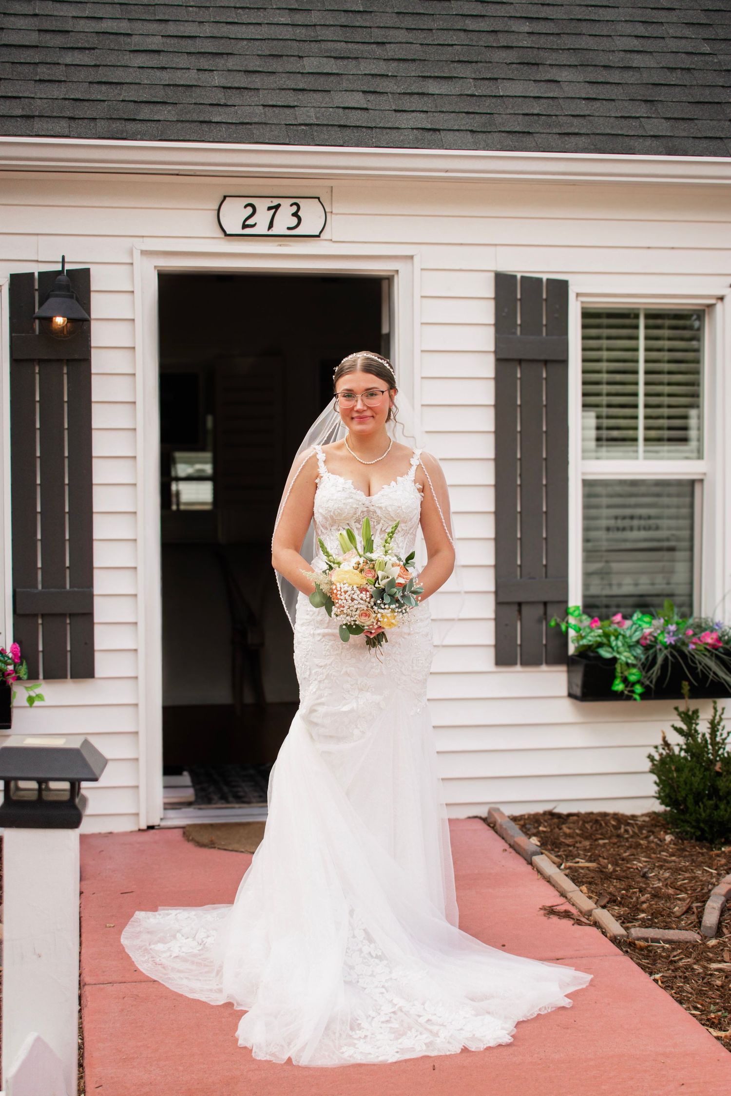 Bride in white lace mermaid gown holding floral bouquet standing in front of house number 273 with dark shutters.