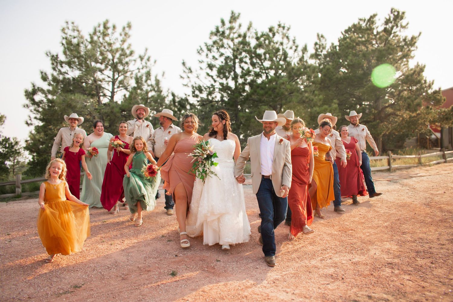 Bride and groom walk with colorful wedding party in mismatched dresses and cowboy hats outdoors.