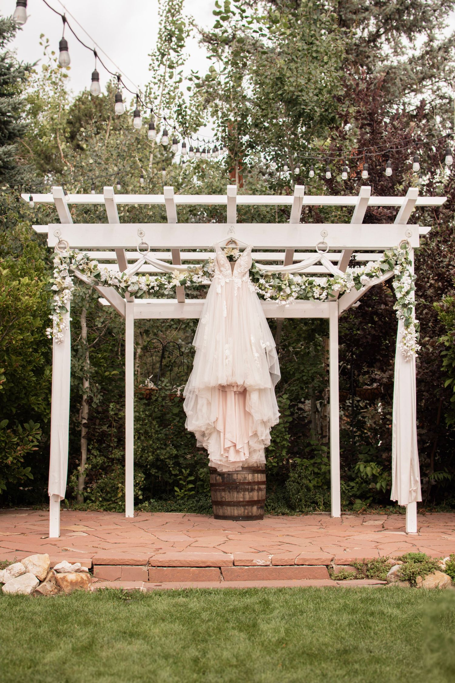 A elegant wedding dress hangs from a white pergola decorated with flowers in a lush garden outdoor ceremony setting.