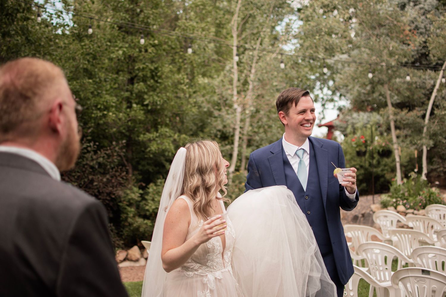 Smiling groom in navy suit walks with bride in veil during outdoor wedding ceremony among trees.