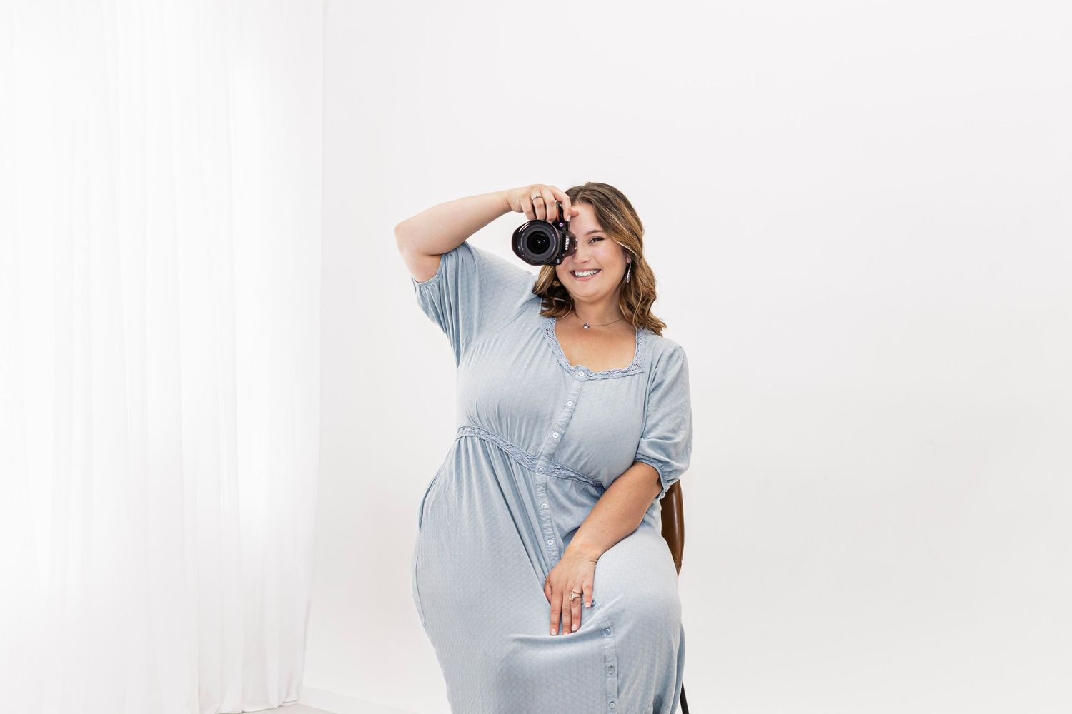 A plus-size woman in a blue dress holds a camera to her eye while posing in a bright white studio setting.