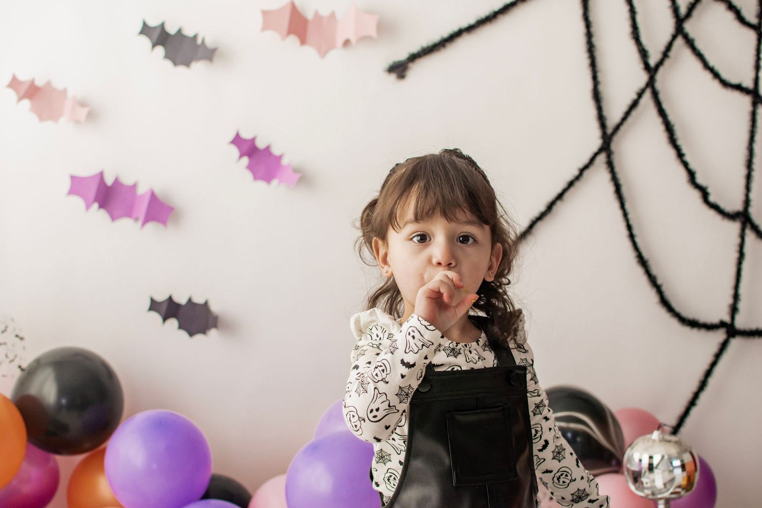 Young girl in Halloween costume sticking out tongue, surrounded by colorful balloons and bat decorations on white wall at Luminous studio