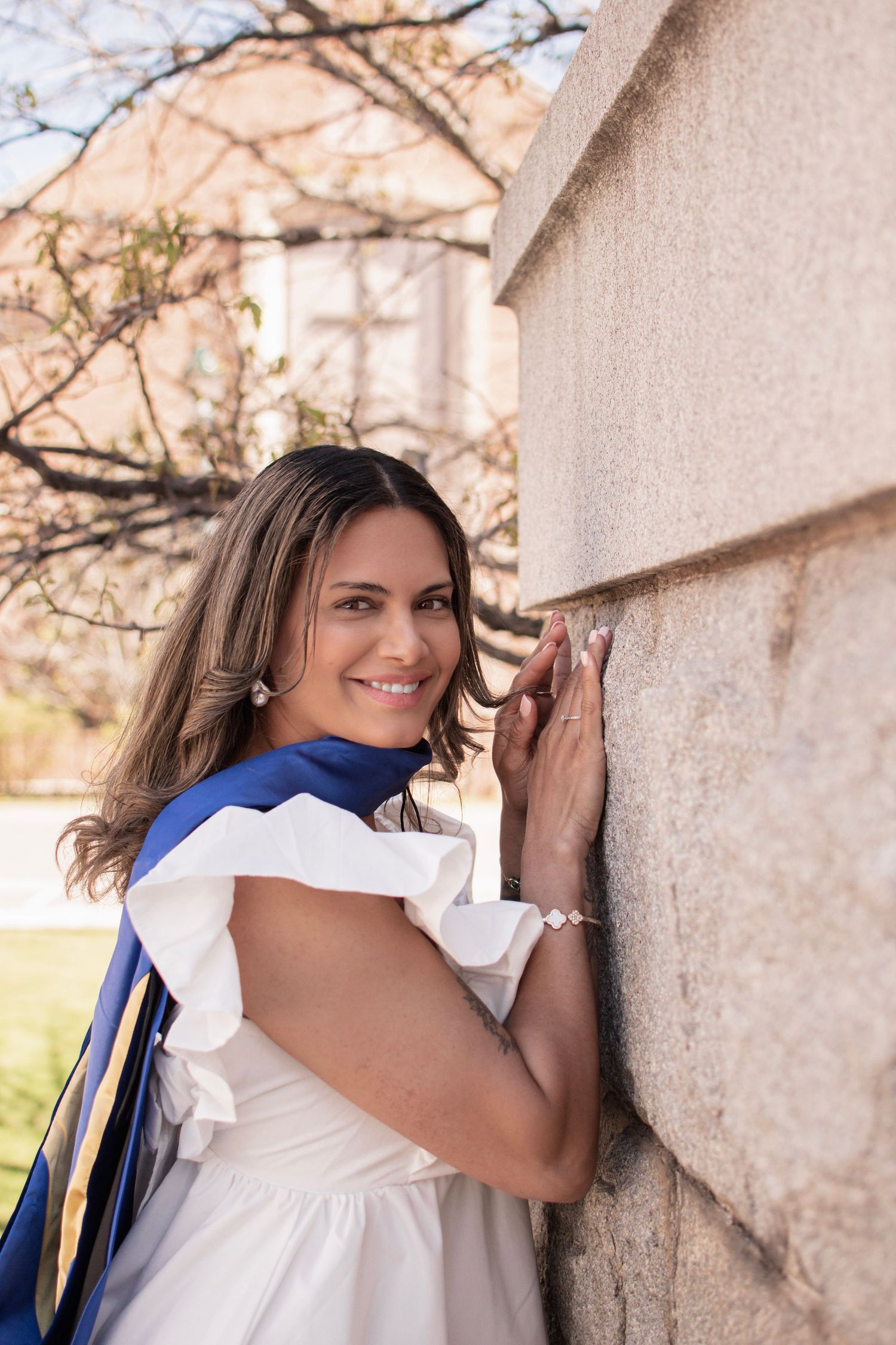 Smiling woman in white dress and blue graduation sash poses against stone wall outdoors on campus.