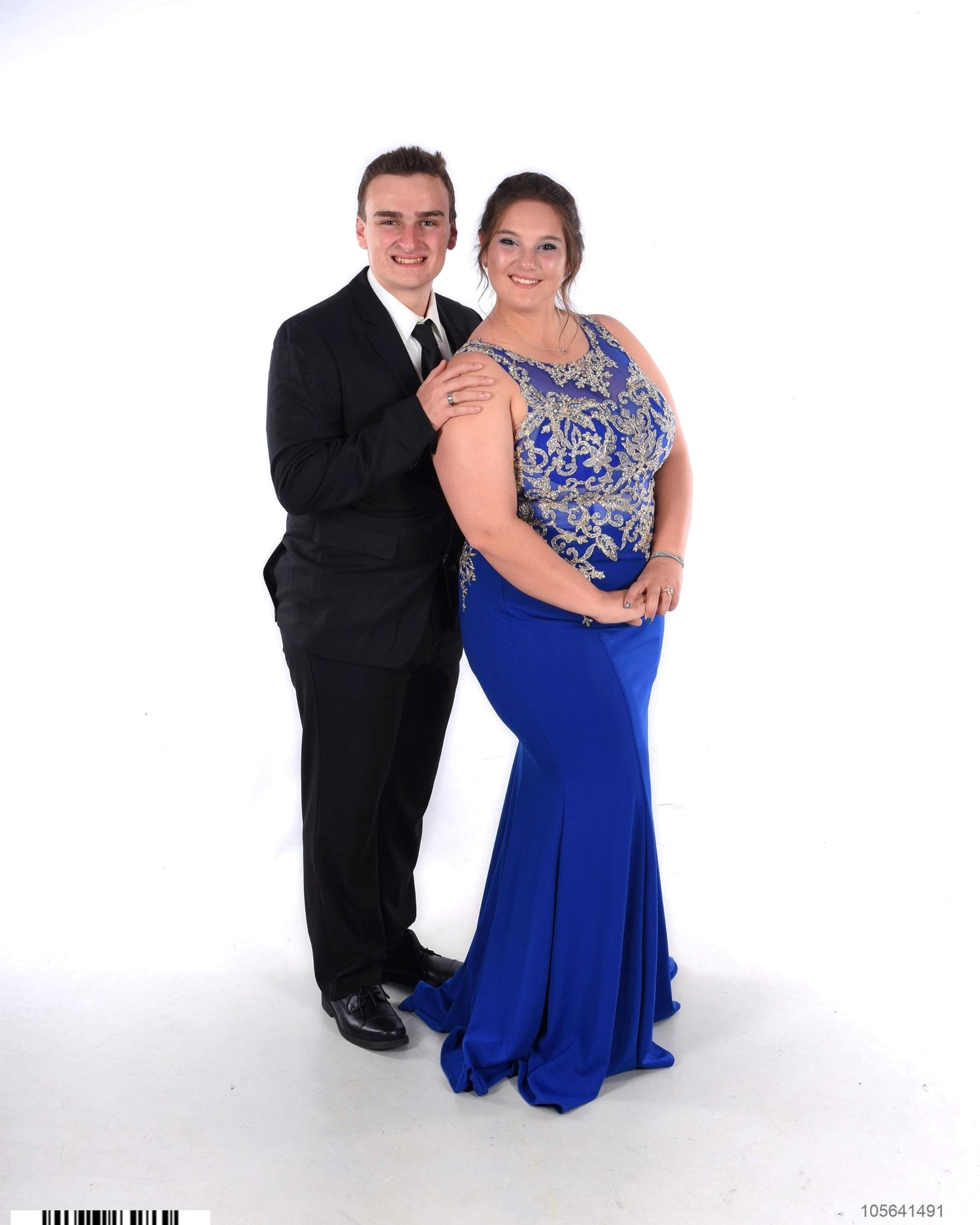 Smiling young couple posing together in formal prom attire, he in a black suit and tie, she in a royal blue beaded mermaid gown.