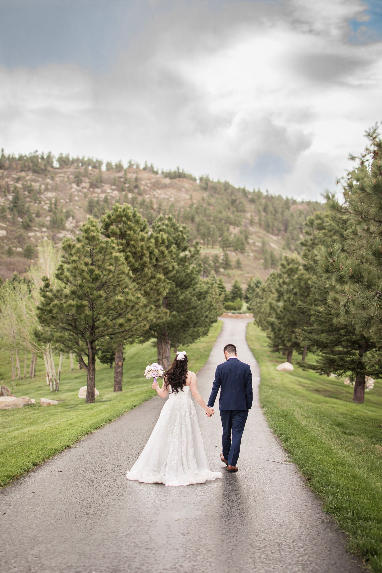 Bride and groom walking hand in hand down a tree-lined road with mountain scenery on their wedding day.
