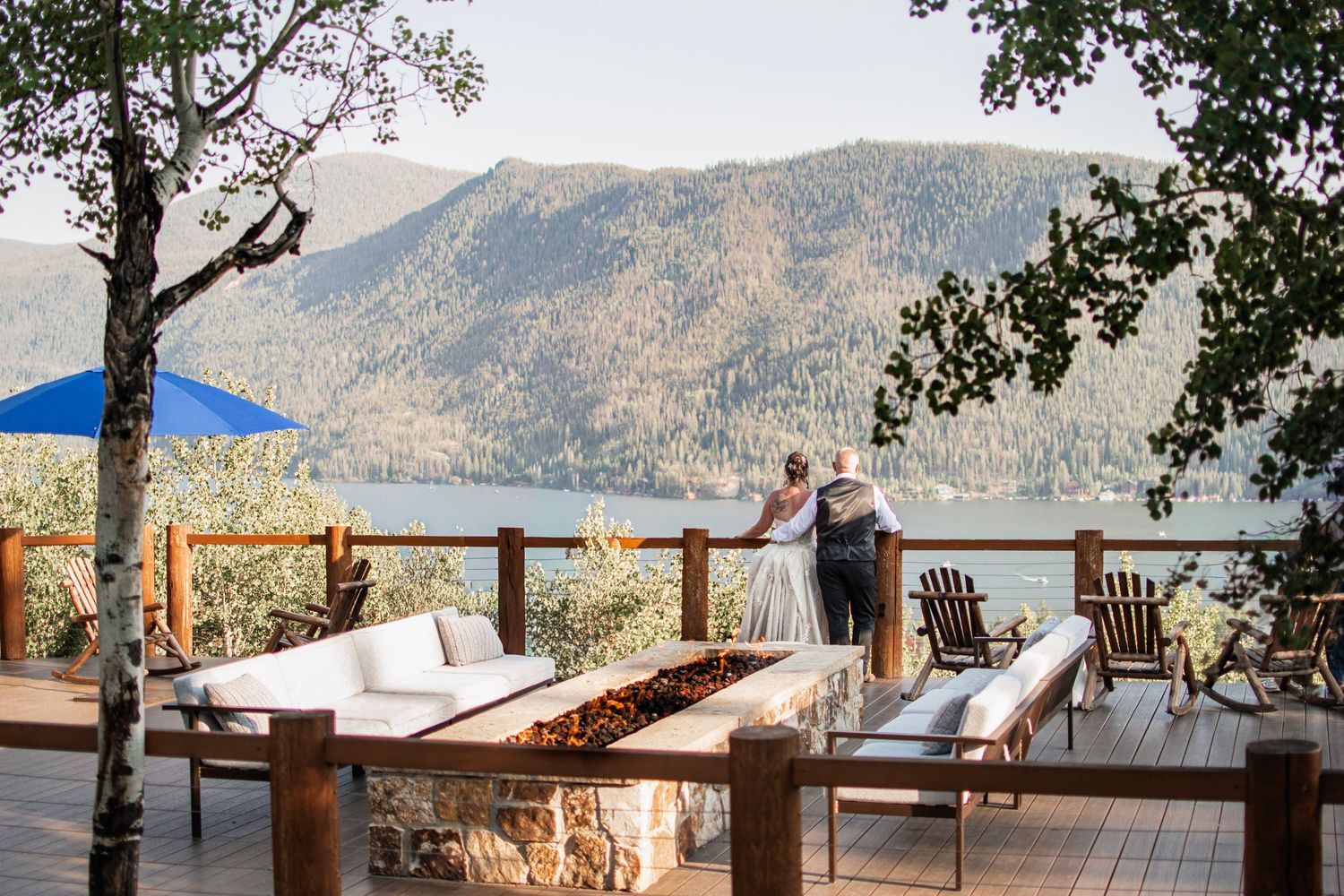 Couple stands on a mountain lodge deck overlooking a scenic lake and forested peaks, with outdoor seating and a stone fire pit.