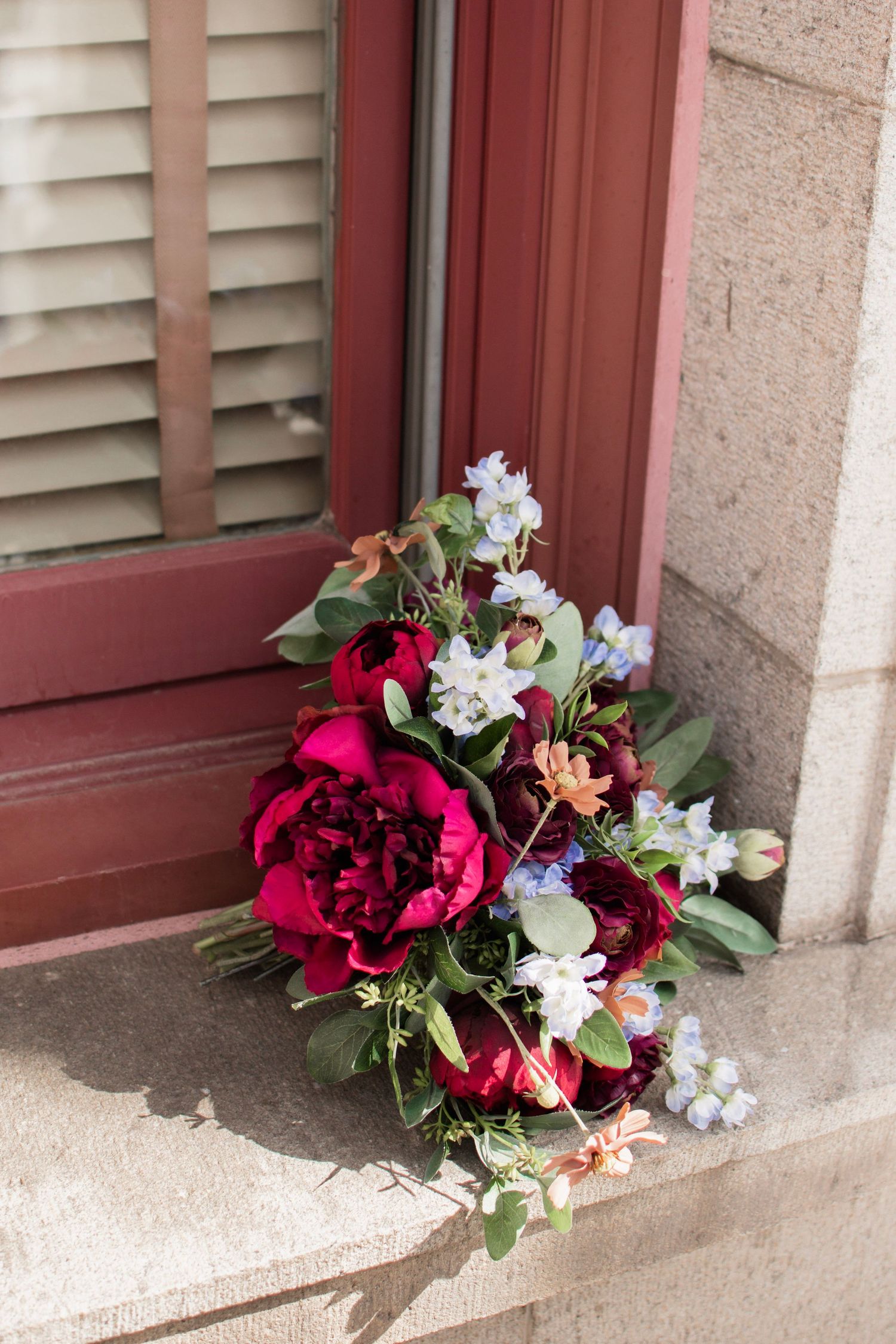 Elegant bridal bouquet with deep red peonies and delicate white flowers resting on a stone windowsill.