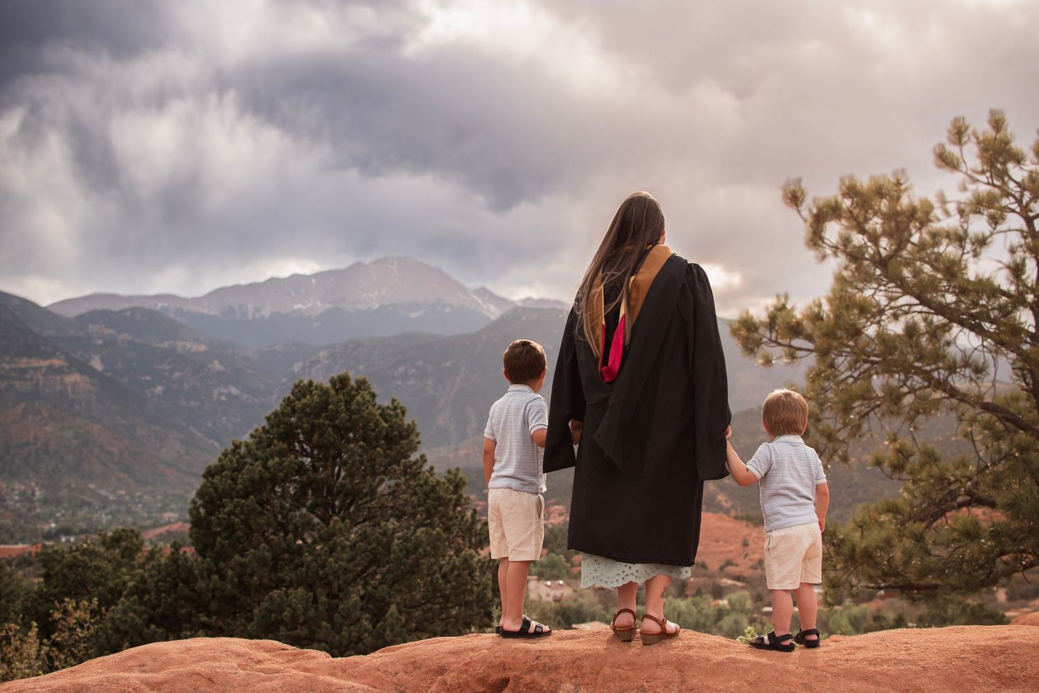 Graduate in black gown holds hands with two young children overlooking a scenic mountain landscape under cloudy skies.