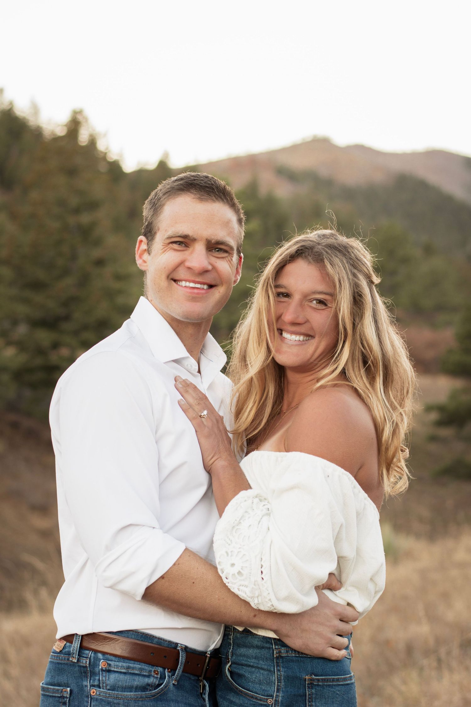 Happy couple embracing outdoors in a mountain meadow, wearing white tops and jeans during golden hour.